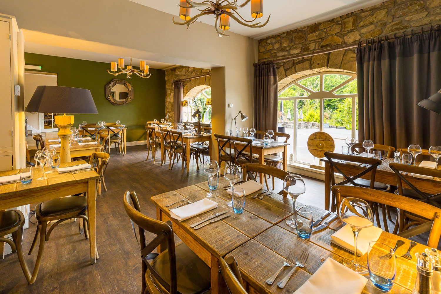 Rustic dining area with wooden tables, stone walls, antler-style chandeliers, and large arched windows overlooking greenery.