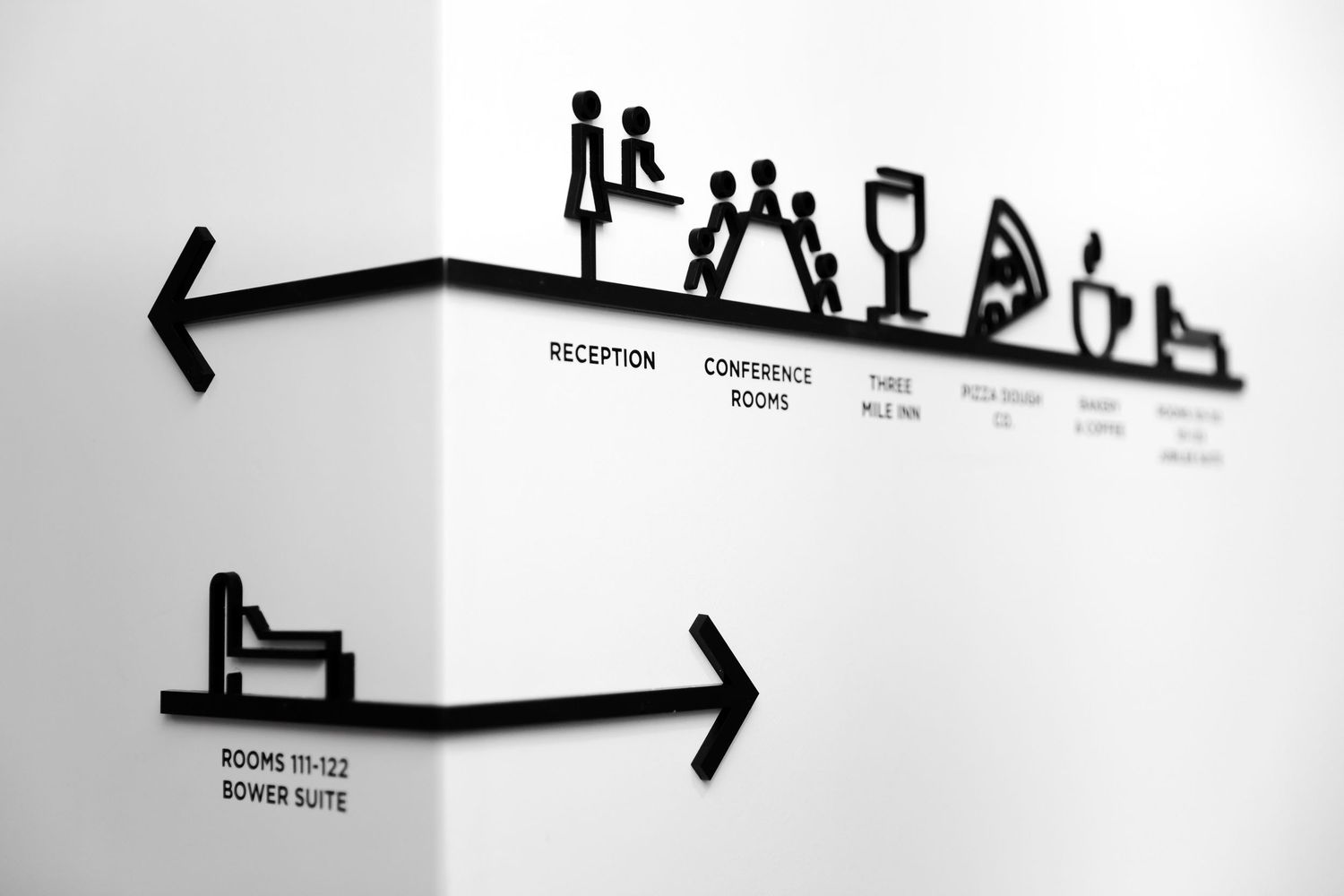 Black directional signage with icons for reception, rooms, dining, and facilities displayed on a clean white corner wall.