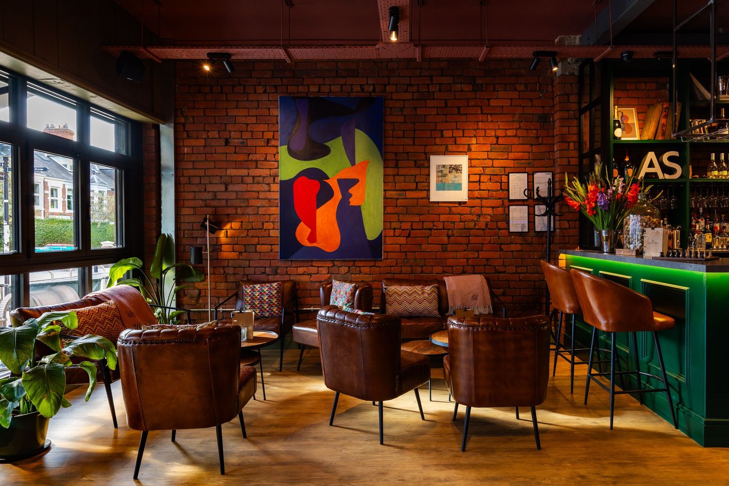 Stylish bar interior with exposed brick walls, leather seating, green bar, and vibrant abstract artwork in warm ambient lighting.