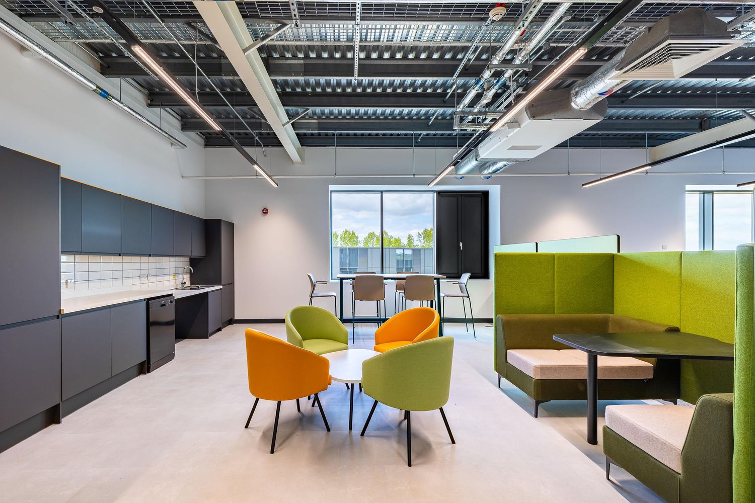 Modern office breakout space with colourful seating, open kitchen area, and green booth pods under exposed industrial-style ceilings.