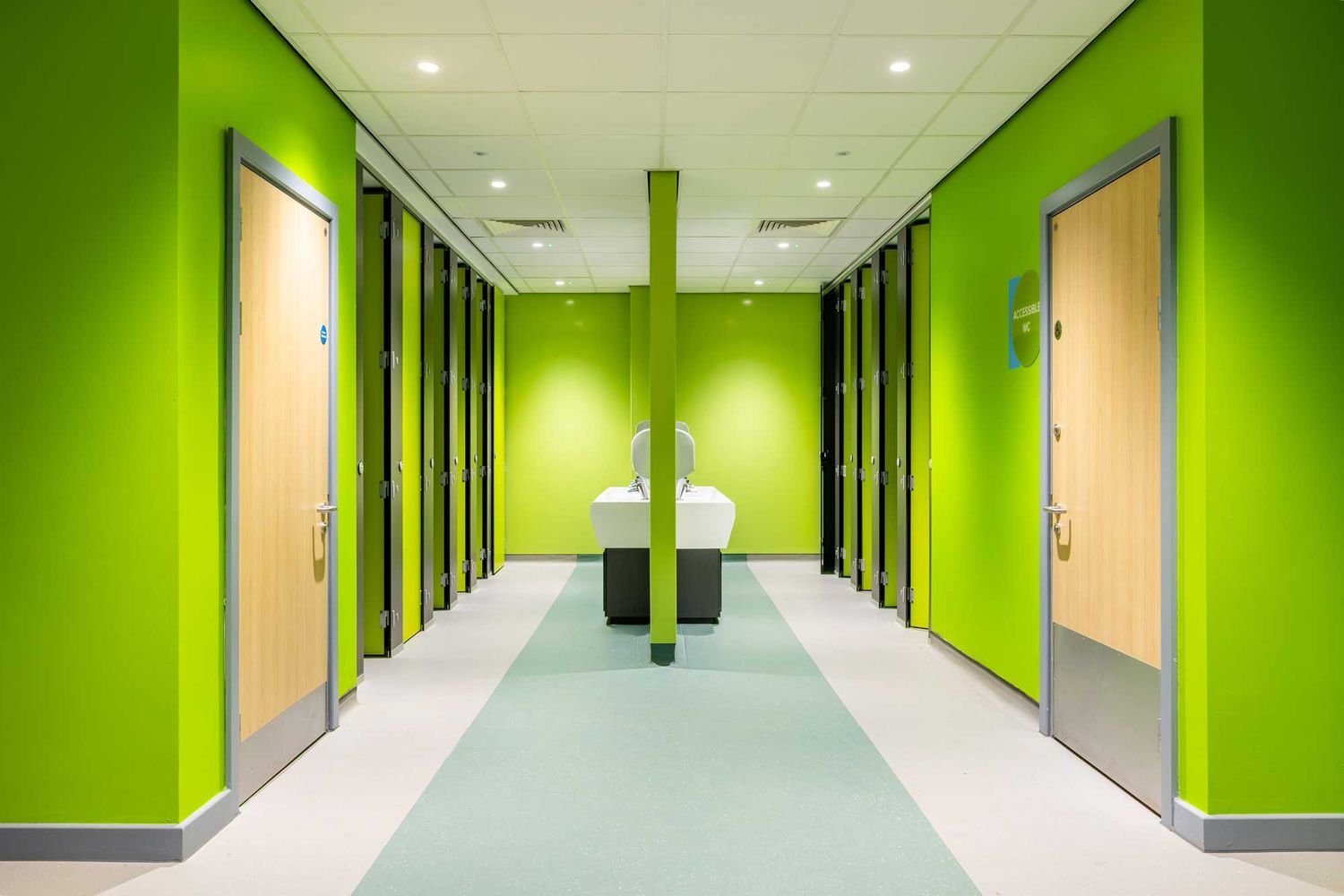 Modern school washroom with bright green walls, symmetrical cubicles & central sinks, showcasing clean lines & contemporary interior design