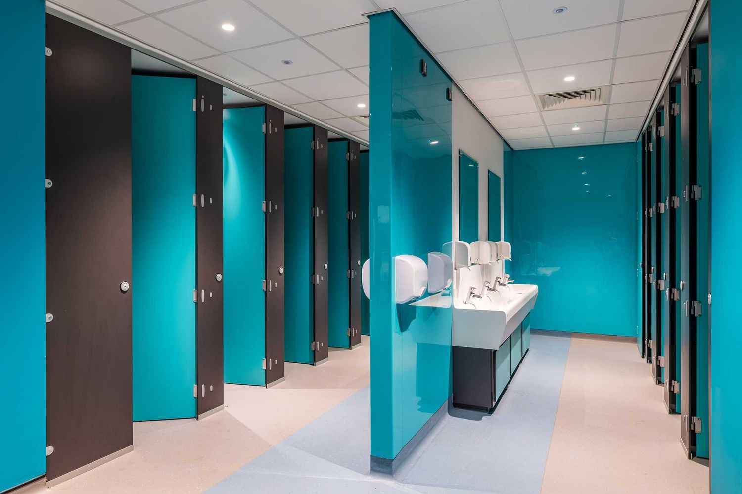 Modern school washroom with turquoise cubicles, clean lines, bright lighting and a central row of sinks in a contemporary interior.