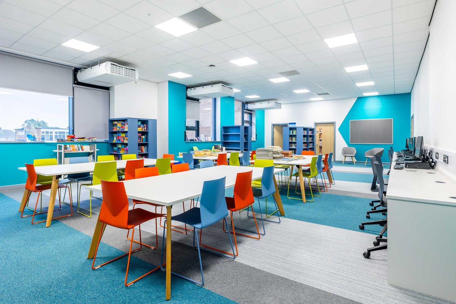 Bright, modern school library with colourful chairs, study tables, bookshelves and computer workstations in a contemporary learning space.