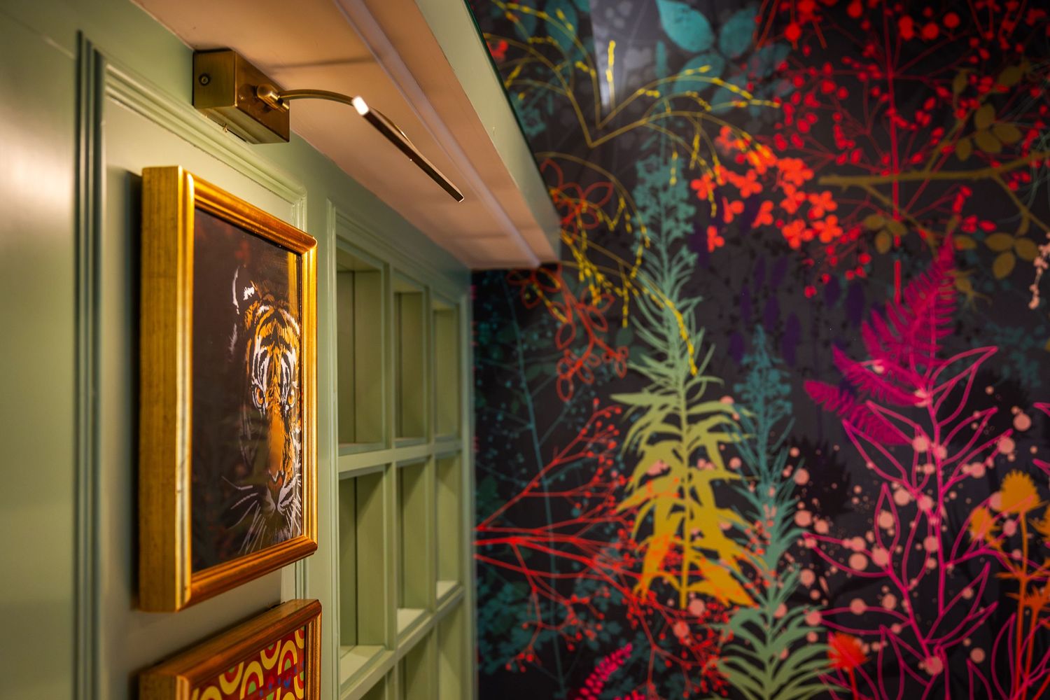 Colourful interior detail featuring a framed tiger portrait, sage green panelling, and vibrant botanical wallpaper.