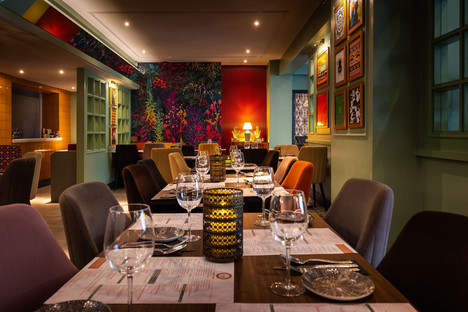 Vibrant dining space with colourful velvet chairs, bold botanical wallpaper, warm lighting, and eclectic framed artwork on green walls.