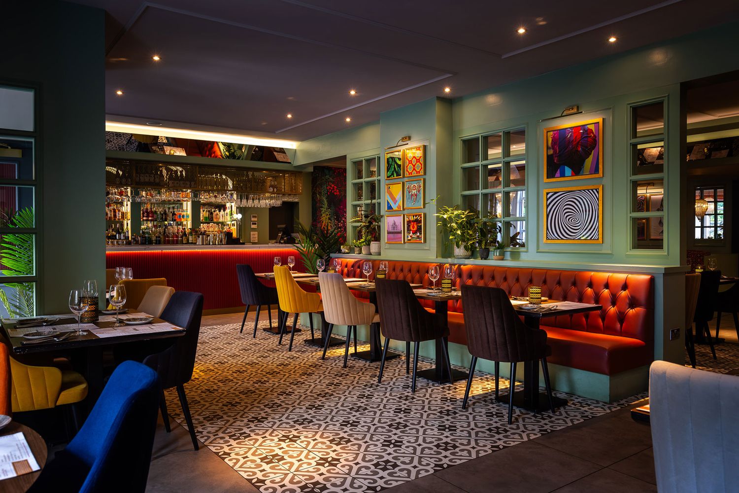 Vibrant restaurant interior with colourful seating, geometric tiled flooring, artistic wall decor, and a warmly lit modern bar area.
