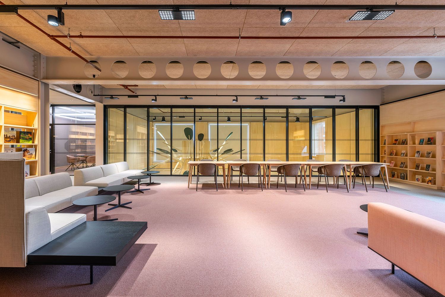 Modern library and collaborative learning space with glass meeting rooms, lounge seating and contemporary furniture in a bright interior.