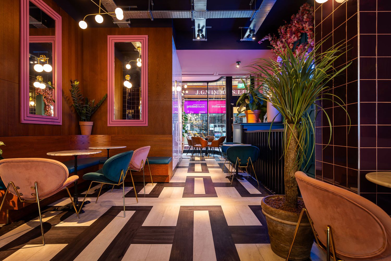 Vibrant café interior with colourful chairs, geometric flooring, pink-framed mirrors, lush greenery, and a cozy, modern ambiance.