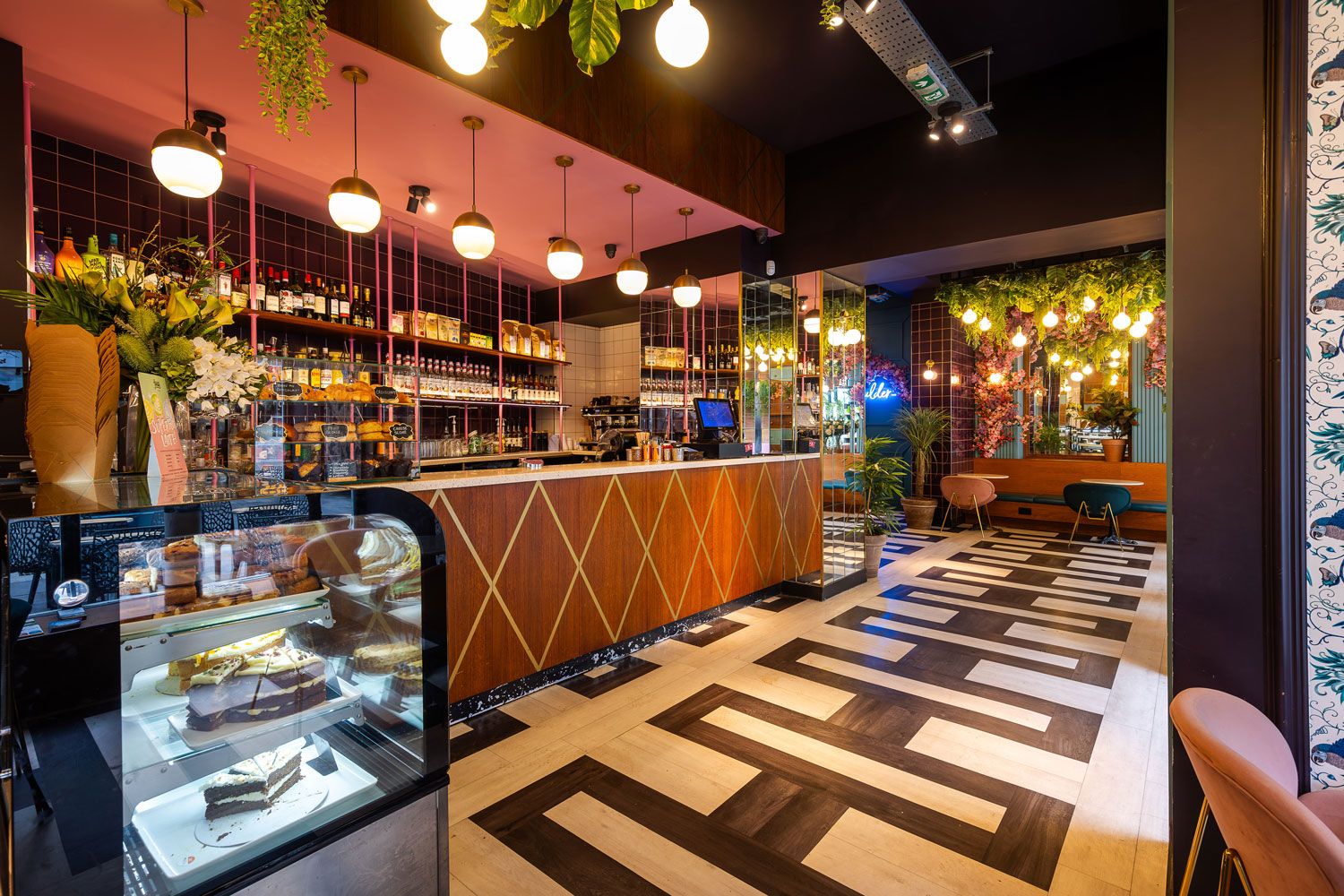 Chic cafe with a geometric wood bar, vibrant pink and gold accents, lush greenery, floral decor, and a stylish tiled black-and-white floor.