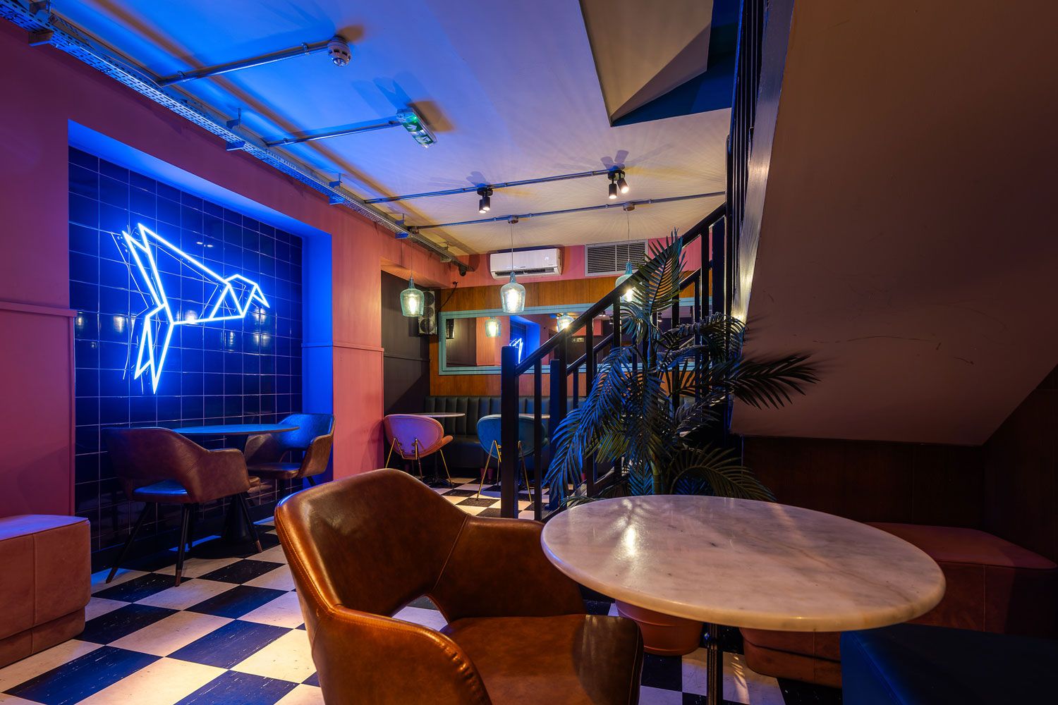 Stylish cafe interior with checkered flooring, vibrant pink walls, neon origami bird art, and cozy seating under warm and modern lighting.