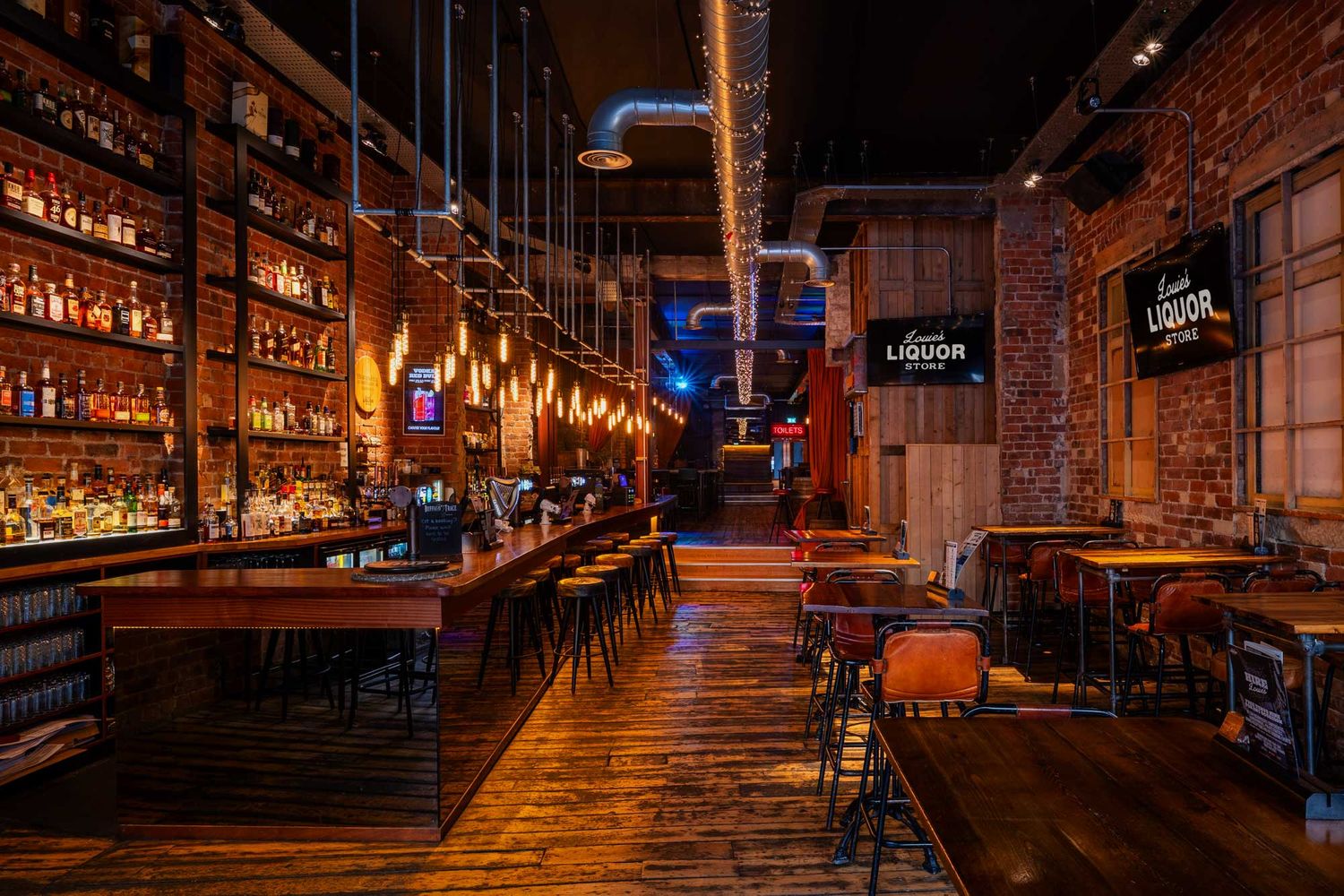 Industrial bar interior with exposed brick walls, wooden floors, hanging lights, and a long stocked bar creating a warm, atmospheric space.