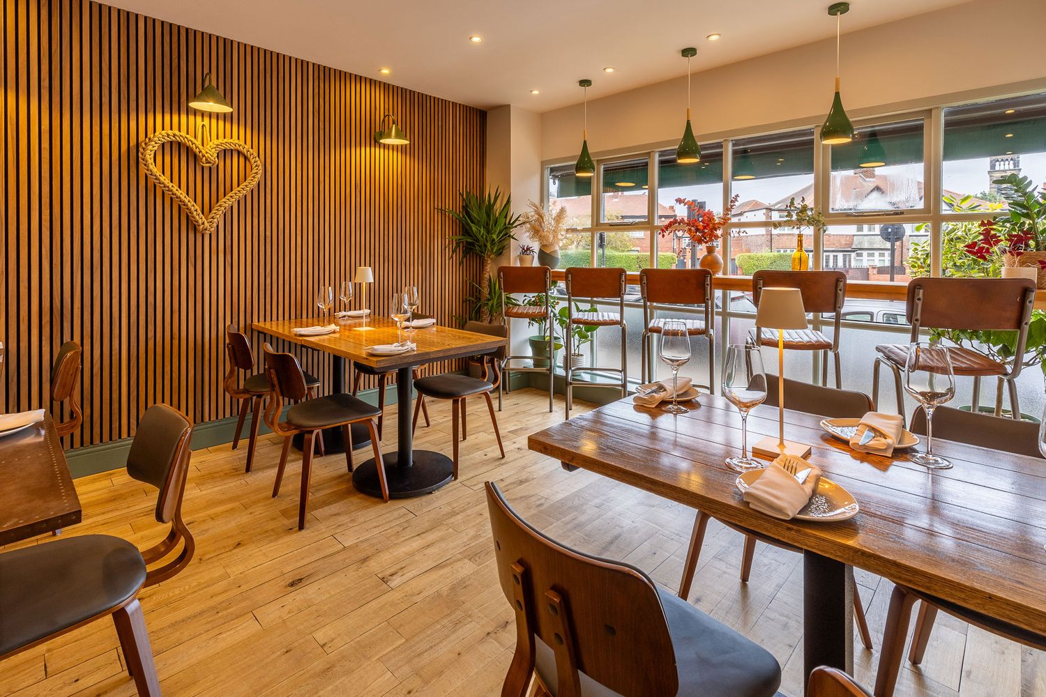 Bright and modern dining space with wood panel walls, large windows, greenery, and a cozy atmosphere featuring elegant table settings.