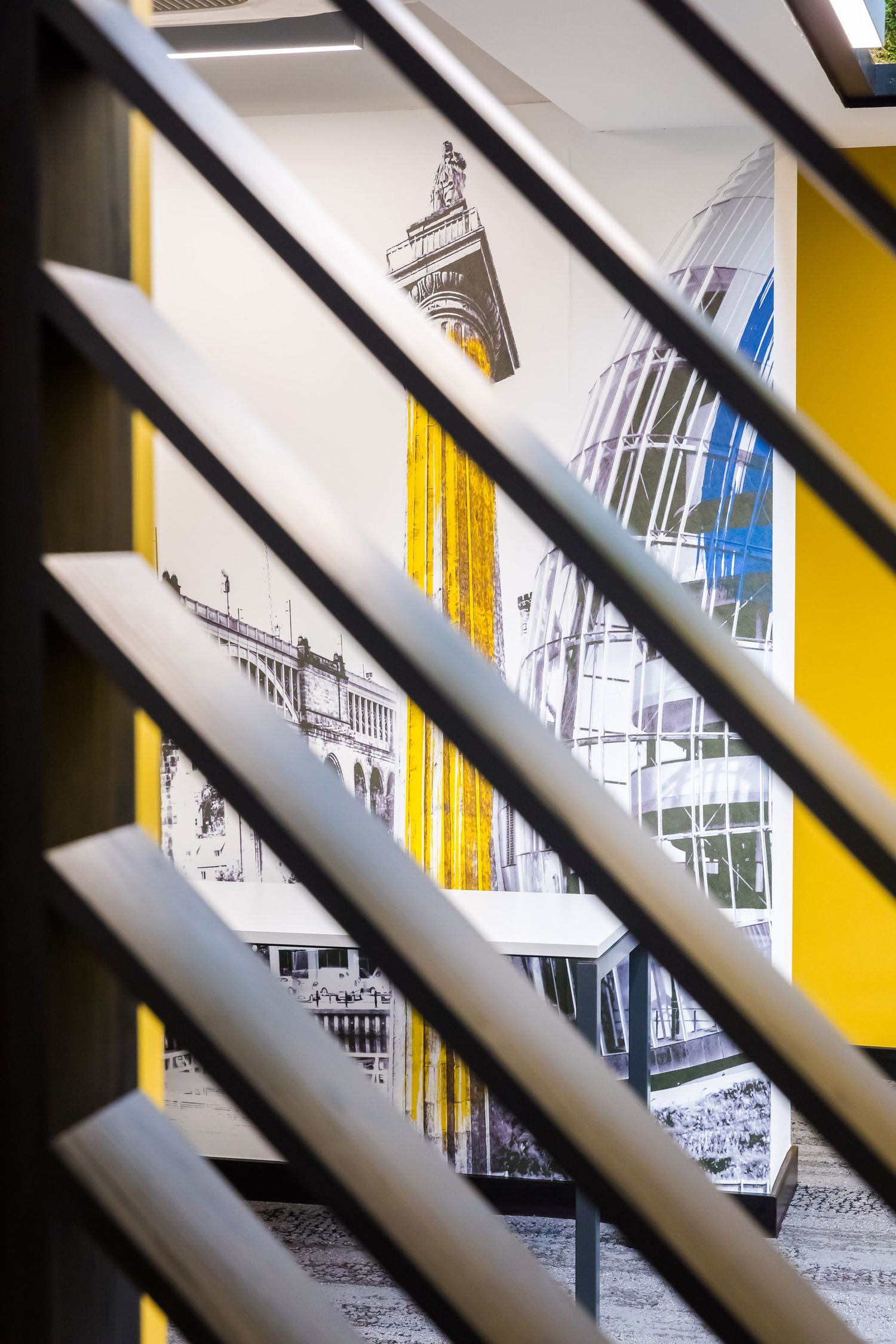 Modern office interior detail with graphic wall art and bold yellow accents, viewed through sleek diagonal stair railings.