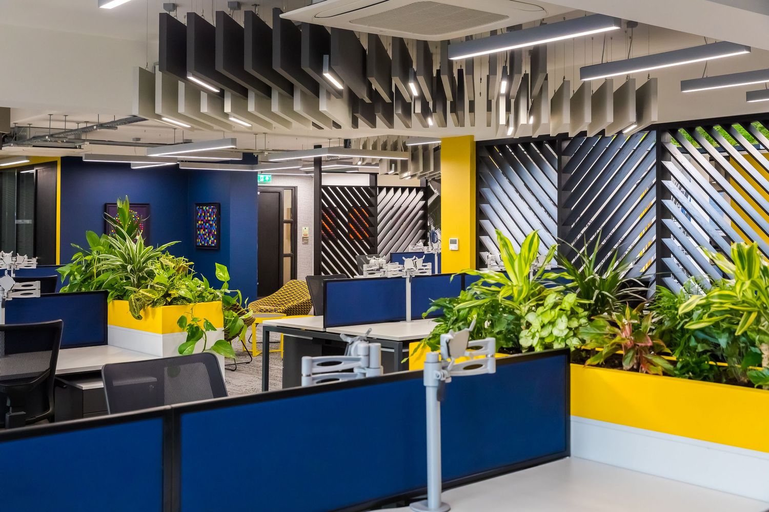 Vibrant open-plan office with bold yellow accents, lush indoor plants, acoustic ceiling panels, and modern workstations.