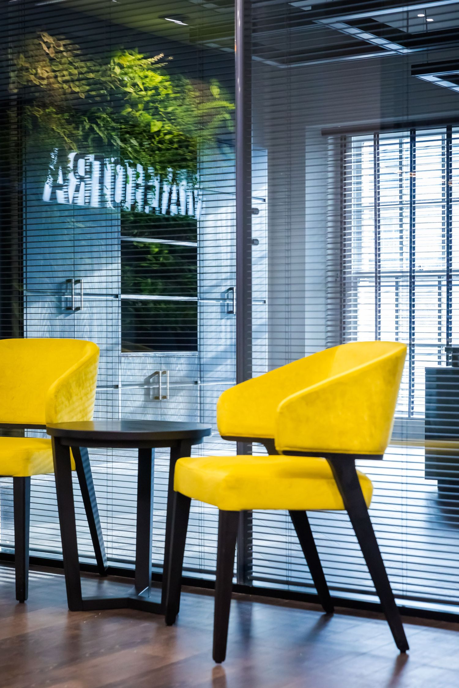 Bright yellow lounge chairs and a small black table set against glass walls with blinds and reflections of greenery in a modern office space