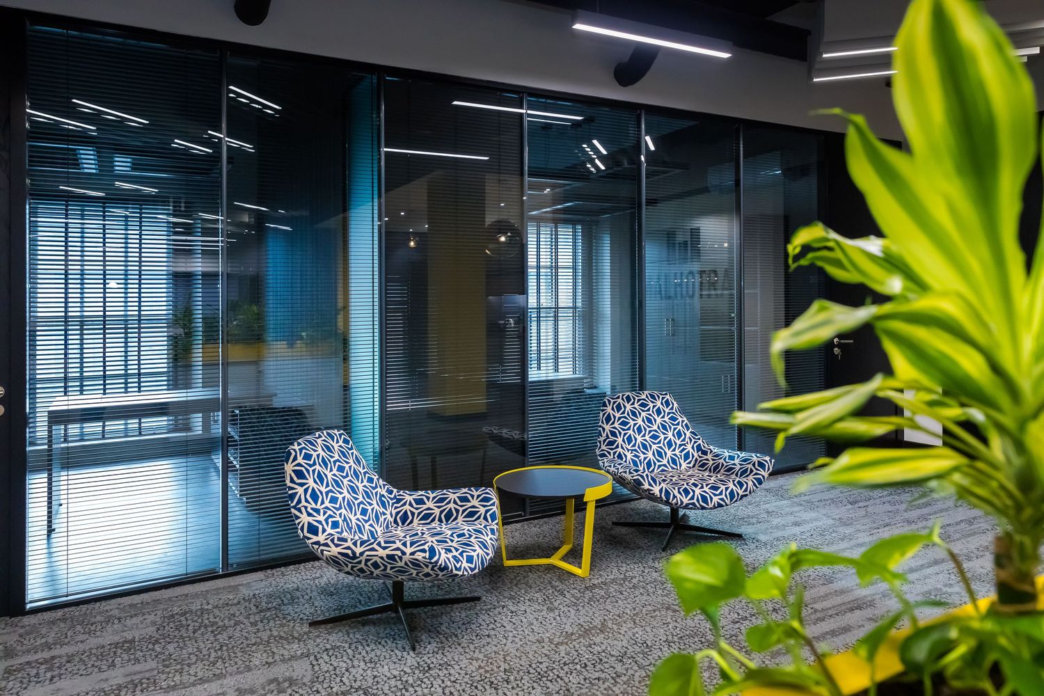 Modern office lounge with patterned chairs, a bold yellow side table, glass partitions, and vibrant greenery adding a fresh, inviting touch.