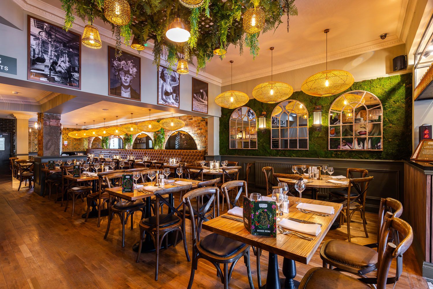 Chic restaurant with wood tables, green moss walls, woven pendant lights, framed mirrors, and artistic black-and-white photography.