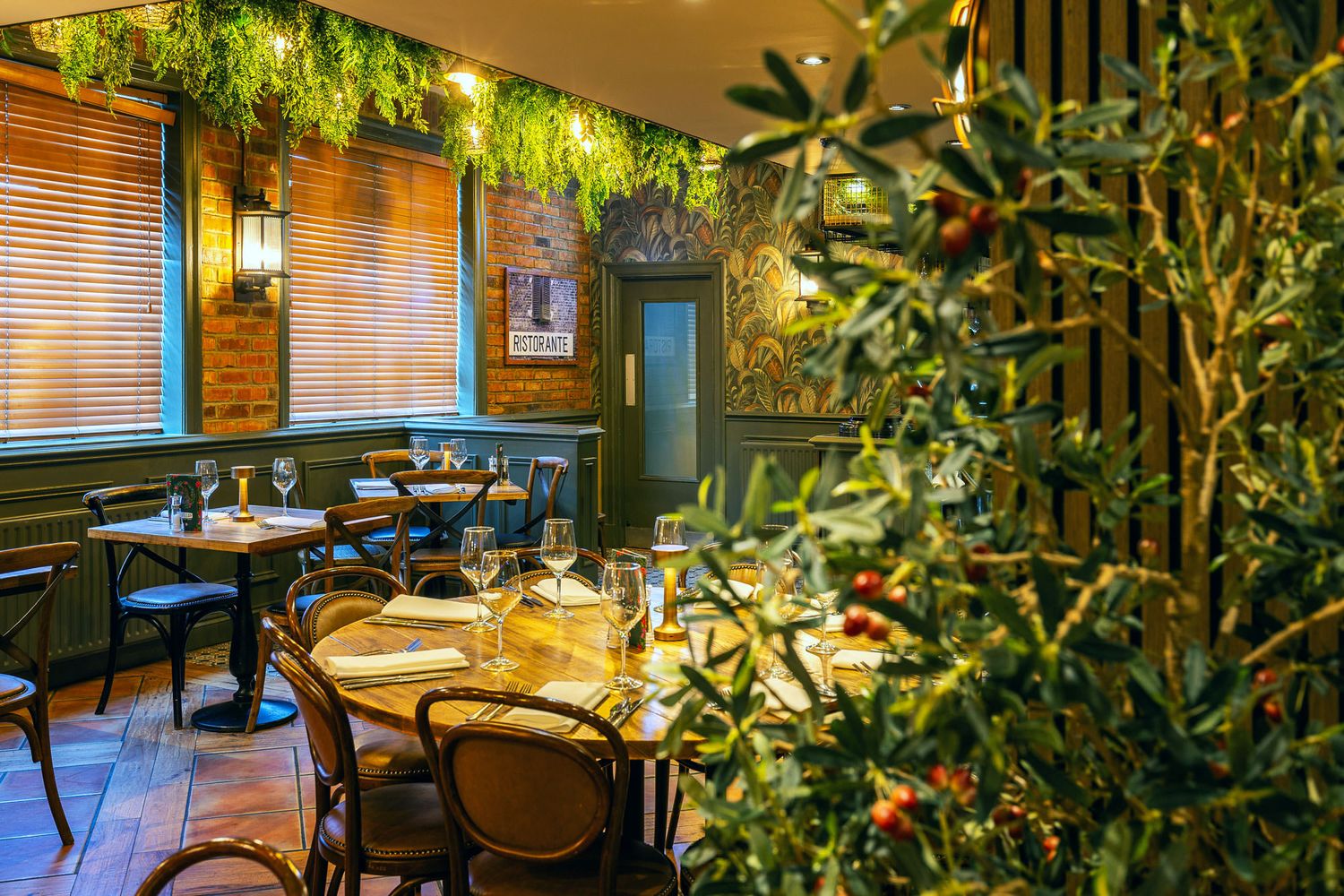 Cozy restaurant with warm wooden tones, lush hanging greenery, patterned wallpaper, and elegantly set tables near brick-accented walls.