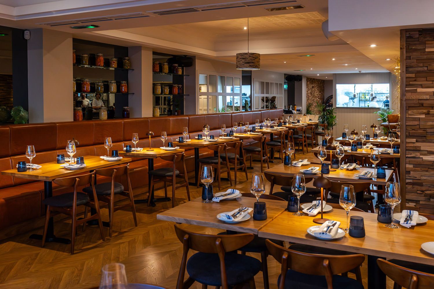 Elegant restaurant interior with wooden tables, leather seating, soft lighting, and jars of preserved ingredients on display.