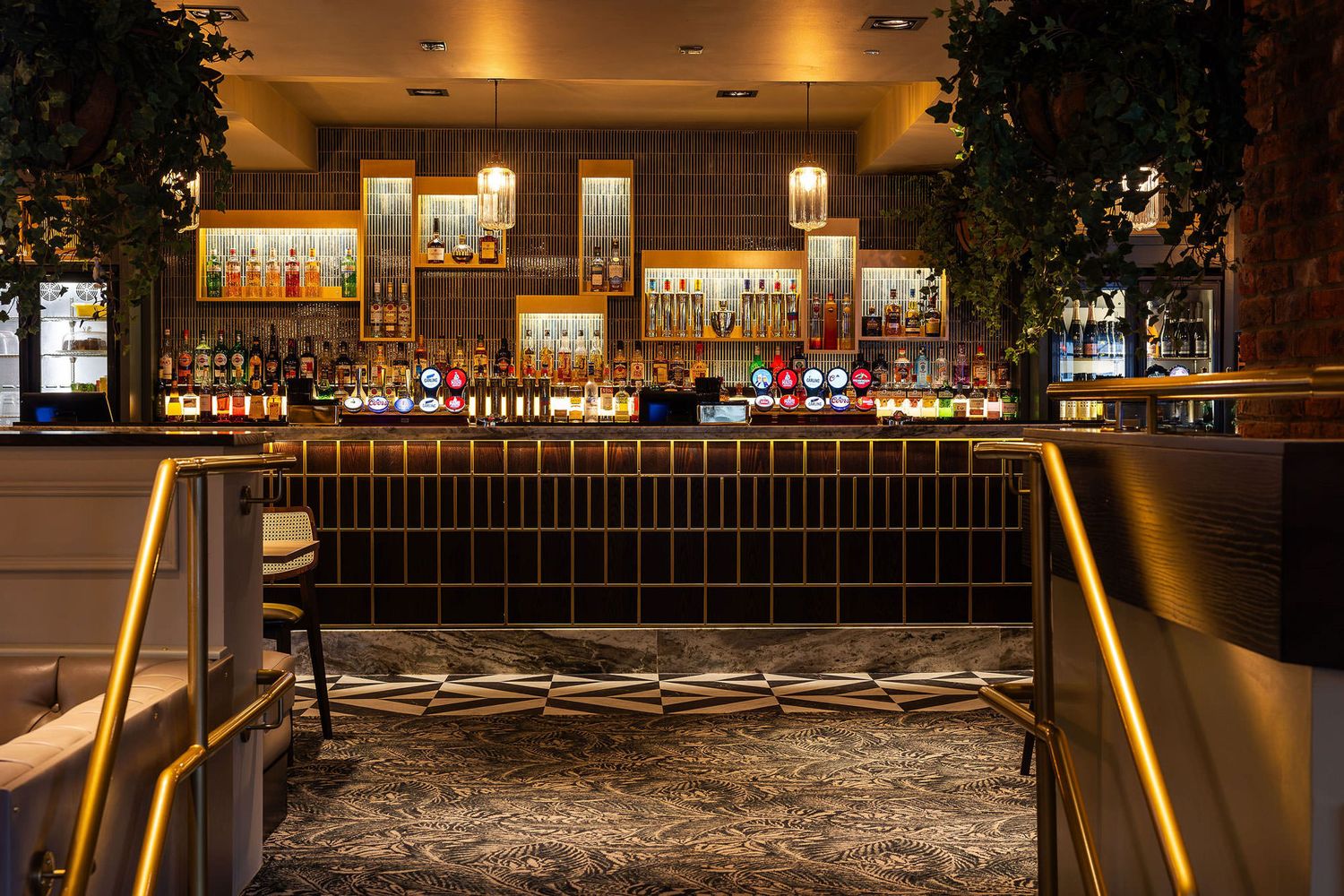 Elegant bar with gold accents, illuminated bottle displays, patterned flooring, and a cozy ambiance with lush greenery and warm lighting.