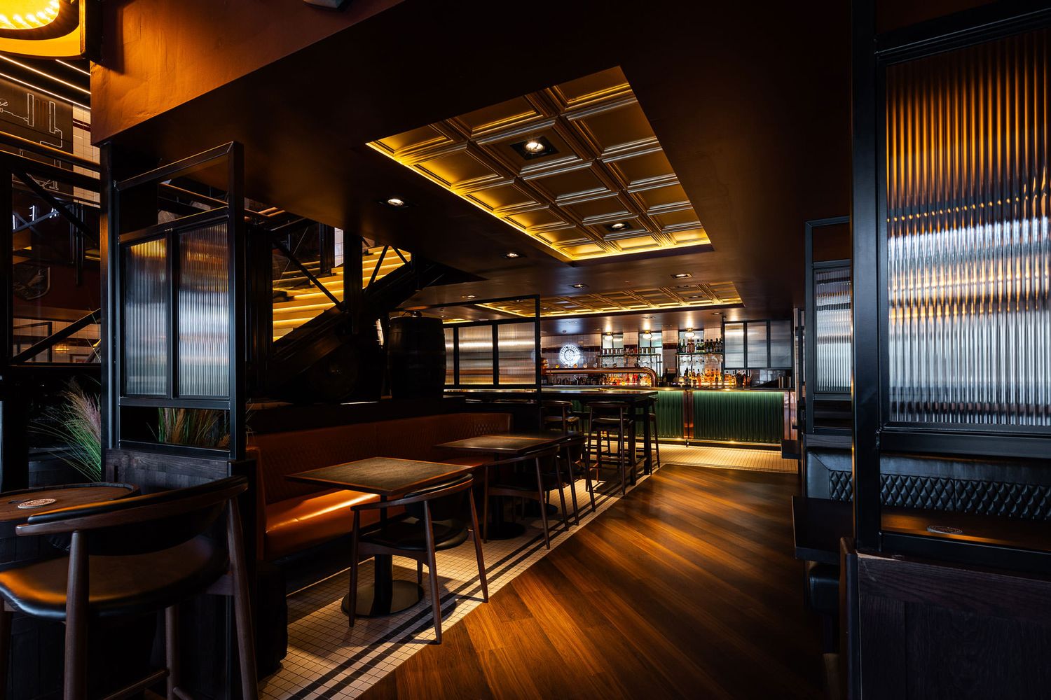 Modern bar interior featuring warm lighting, wood panelled floors, green bar counter, textured glass partitions, and cozy leather seating.
