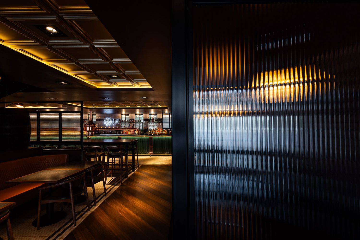 Moody bar interior with dark wood floors, textured glass partitions, warm lighting, and a sophisticated ceiling design.