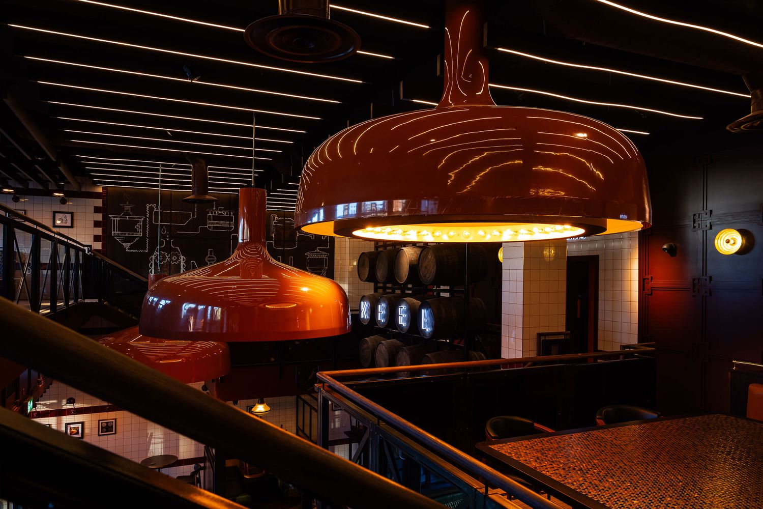 Industrial-style brewery interior featuring large orange brewing tanks, barrel decor, dark tones, and warm lighting accents.