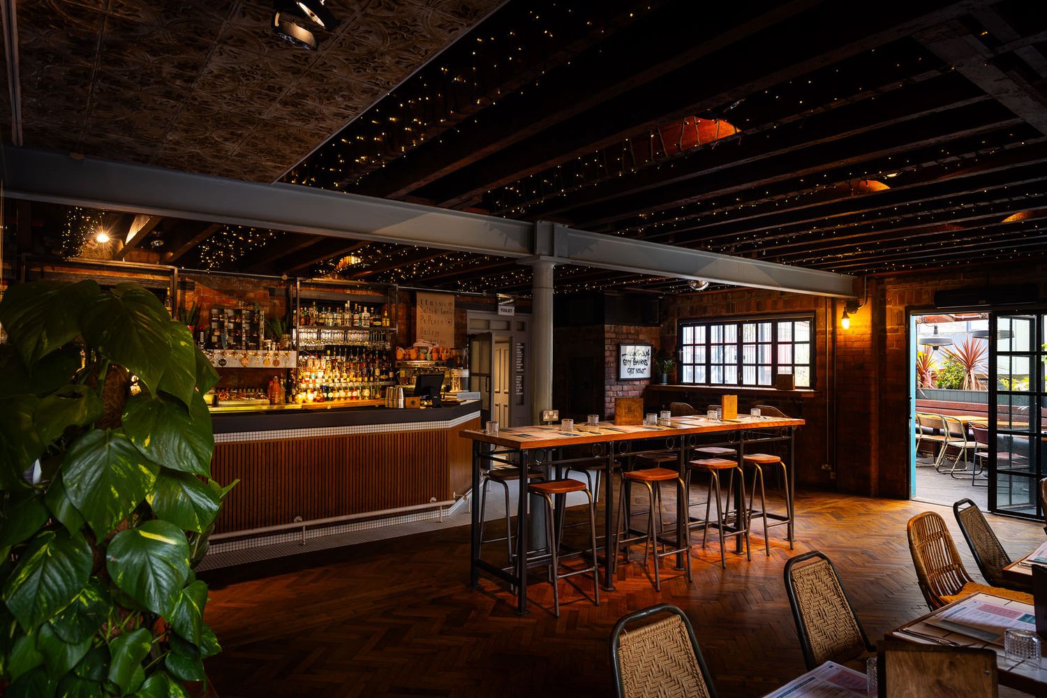 Rustic bar with exposed beams, warm lighting, wood accents, a lush plant centrepiece, and a cozy outdoor seating area in view.