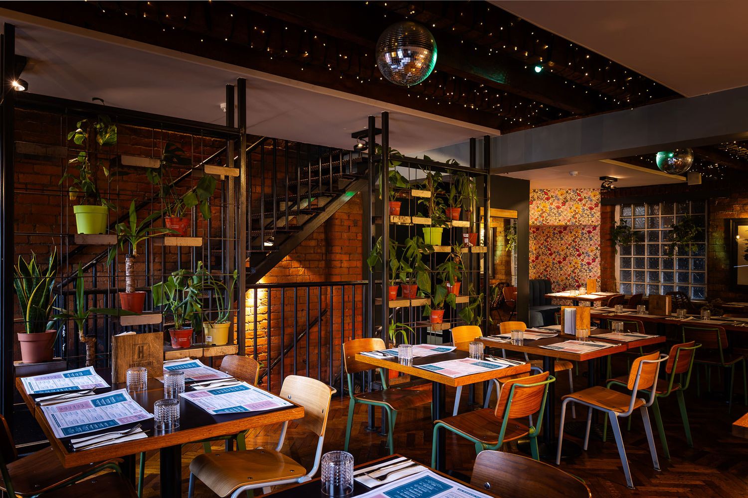 Cozy dining space with industrial shelving, potted plants, exposed brick walls, string lights, and a staircase with a disco ball overhead.