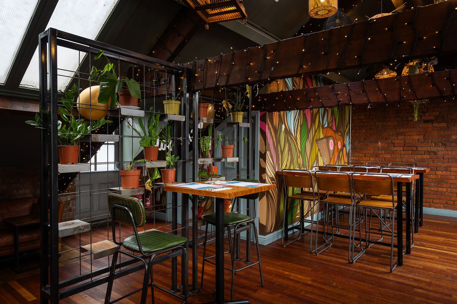 Cafe with potted plants, colourful mural, exposed brick, wood floors, skylights, and industrial-style seating under warm string lights.