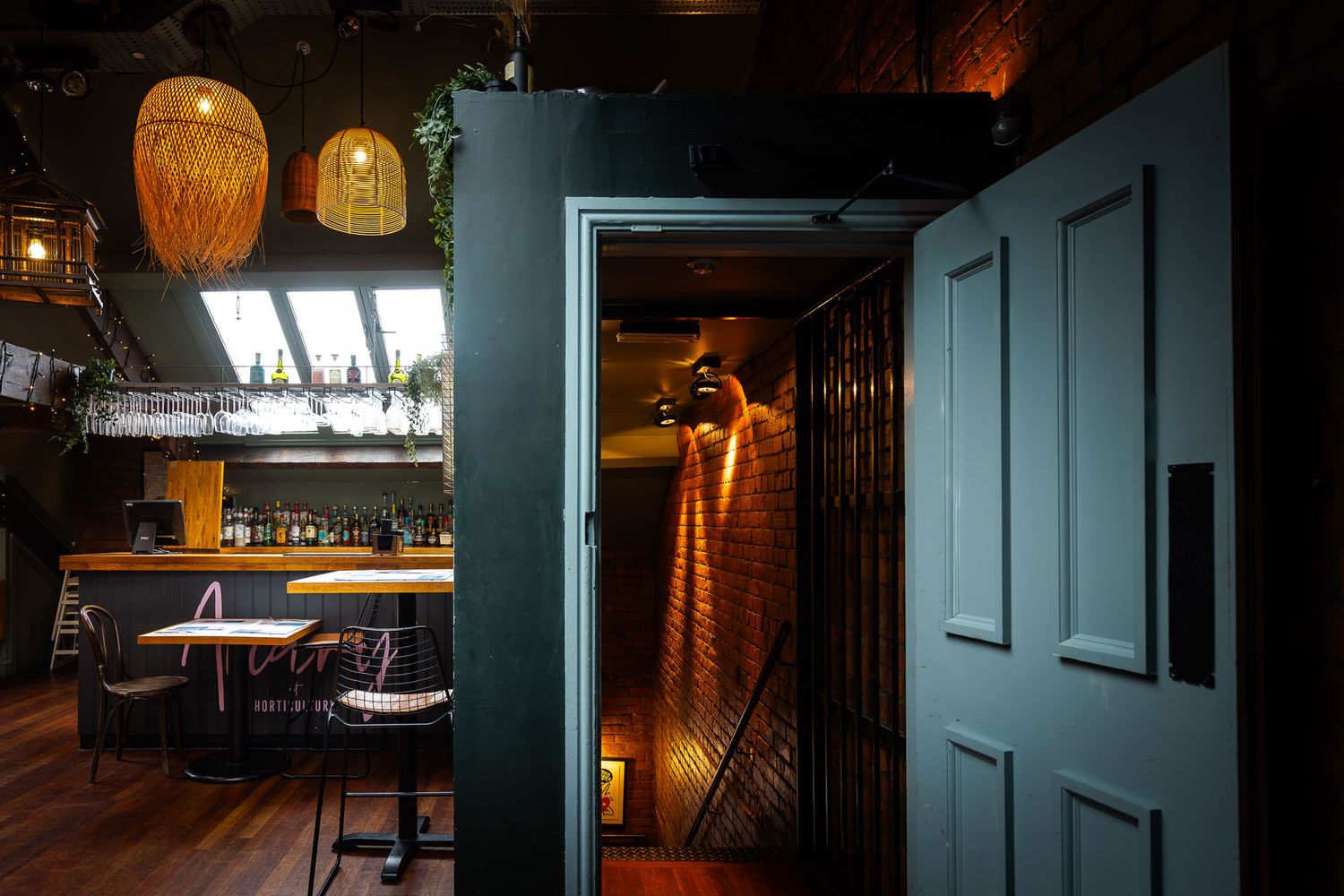 Modern bar with skylights, hanging wicker lamps, a vibrant green wall, and an inviting staircase leading to a cozy brick-walled lower level.
