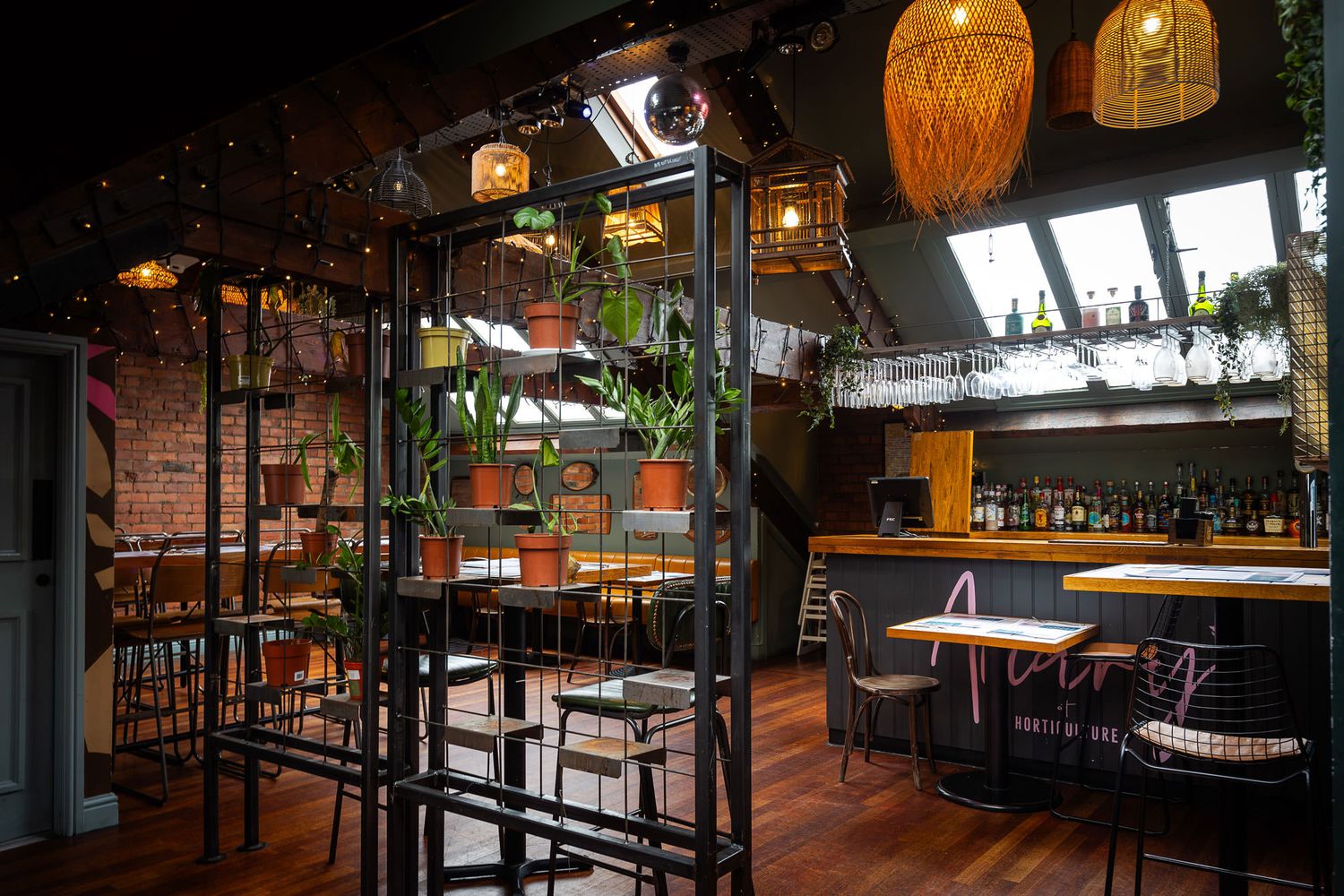Eclectic bar and dining area with hanging lights, potted plants, skylights, and a vibrant mix of industrial and natural decor elements.