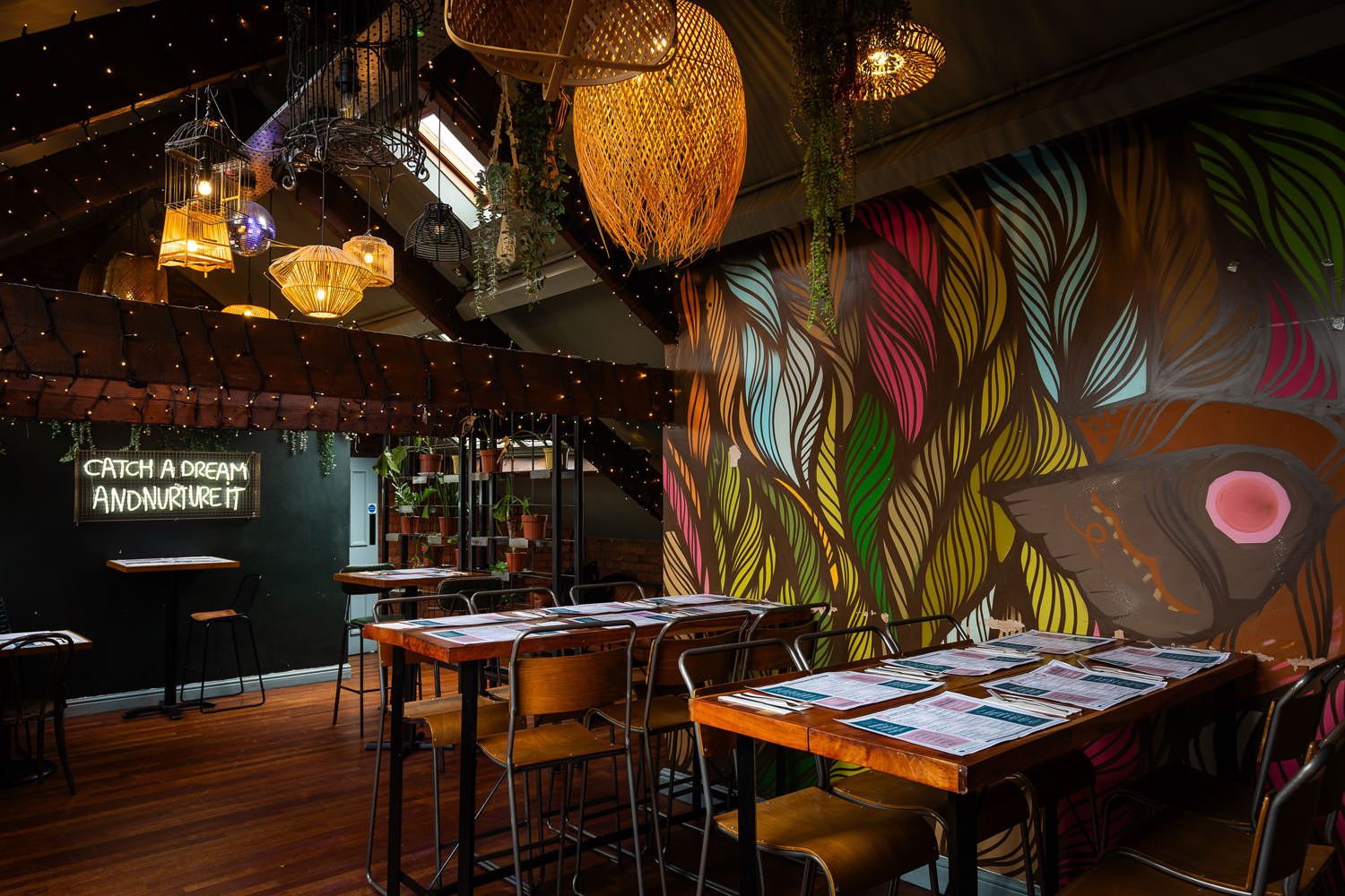 Eclectic dining area with vibrant mural, string lights, hanging wicker lamps, and a neon sign that reads 'Catch a dream and nurture it.