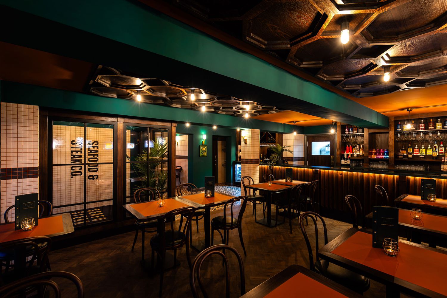 Stylish seafood and steak restaurant with teal walls, warm lighting, tiled accents, wood details, and a geometric ceiling design.