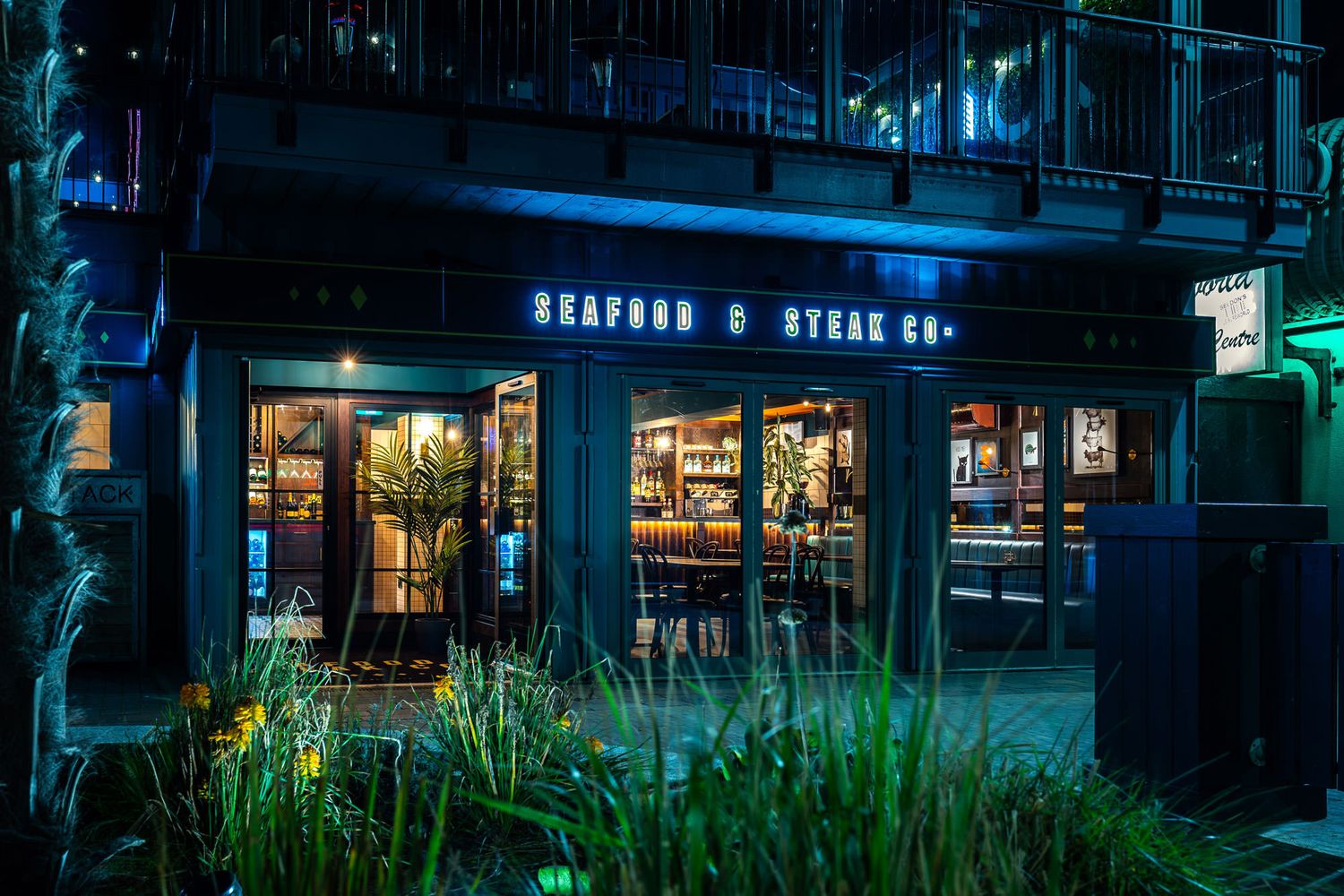 Nighttime view of Seafood & Steak Co. with glowing neon signage, glass doors, modern decor, and lush greenery in the foreground.