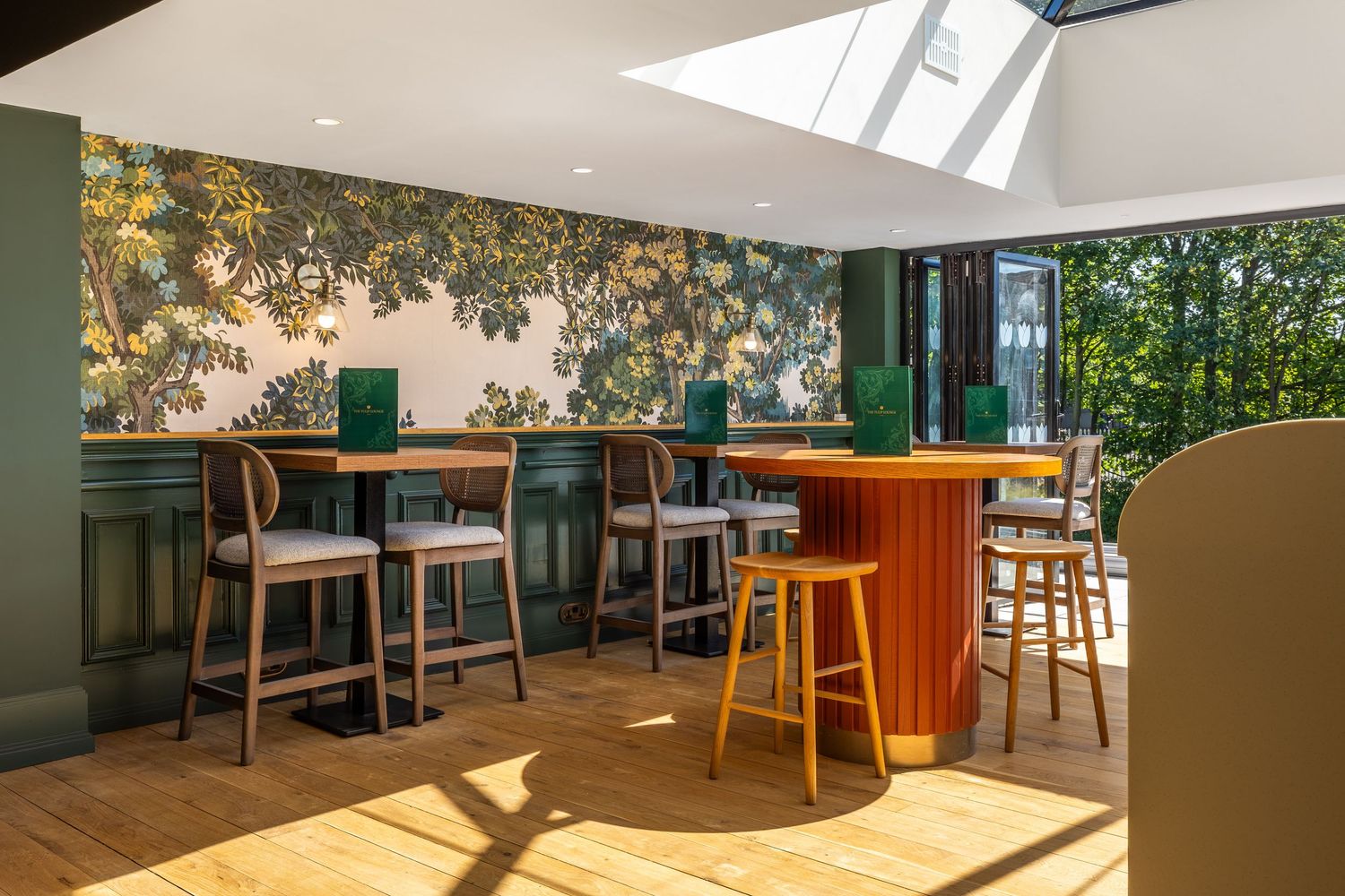 Bright café interior with botanical wallpaper, green paneling, and wooden furniture bathed in natural light from large windows.