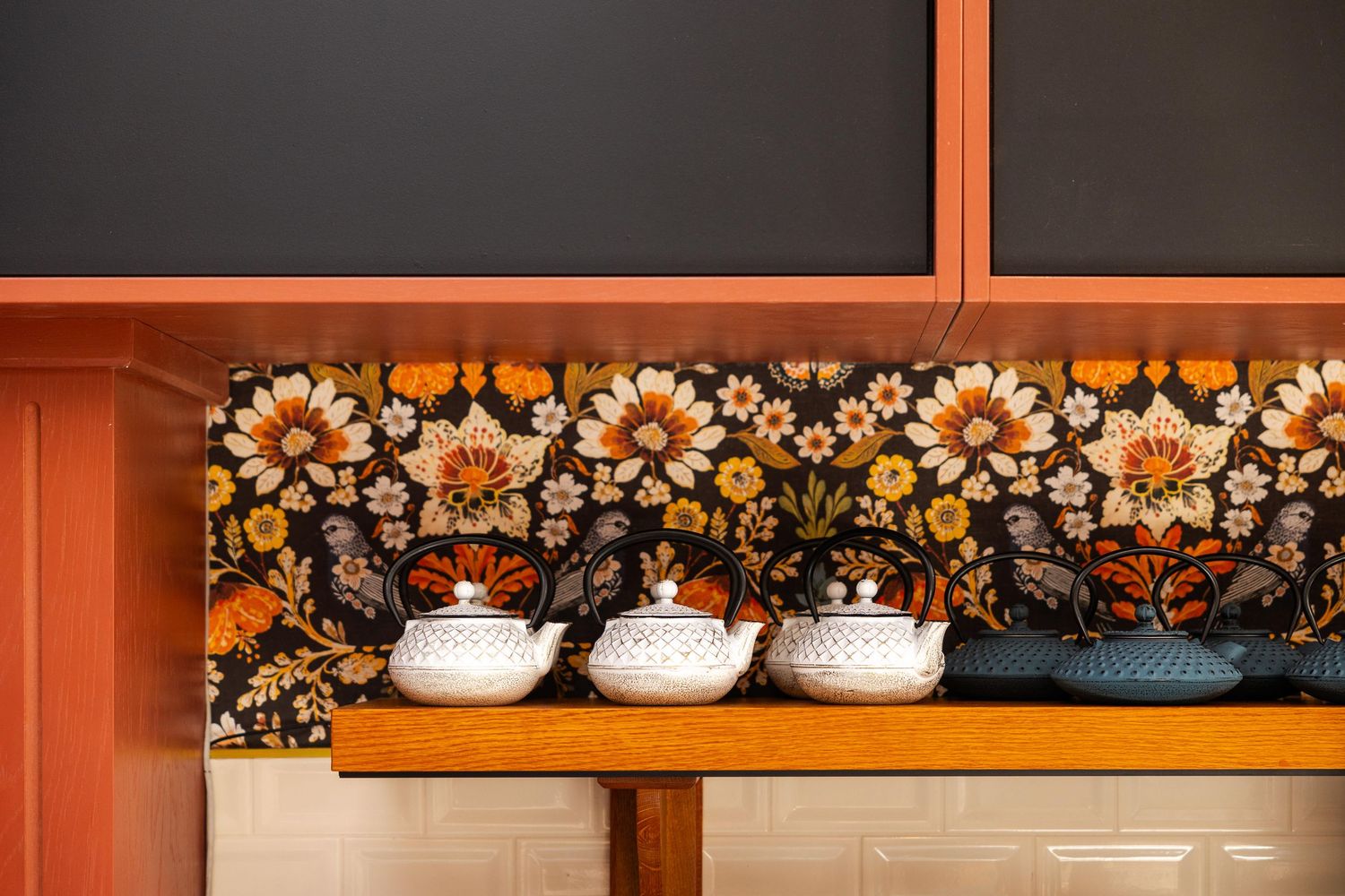 Close-up of teapots on a wooden shelf against a bold floral backsplash and warm terracotta cabinetry.