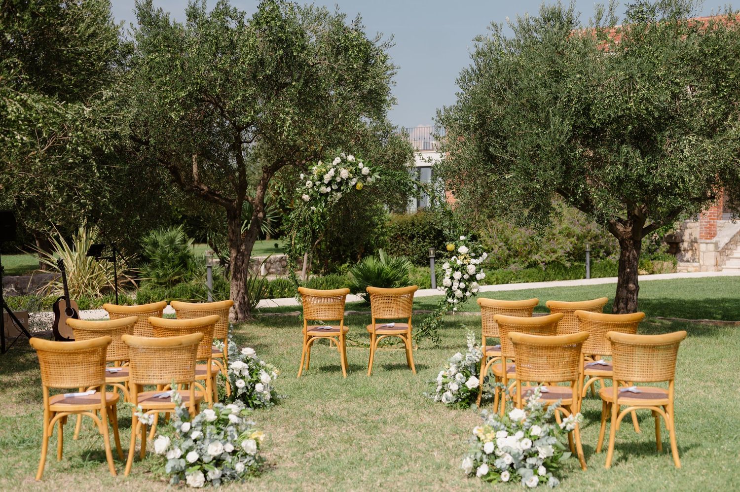 Garden wedding ceremony setup with wooden chairs and white floral arrangements among olive trees.