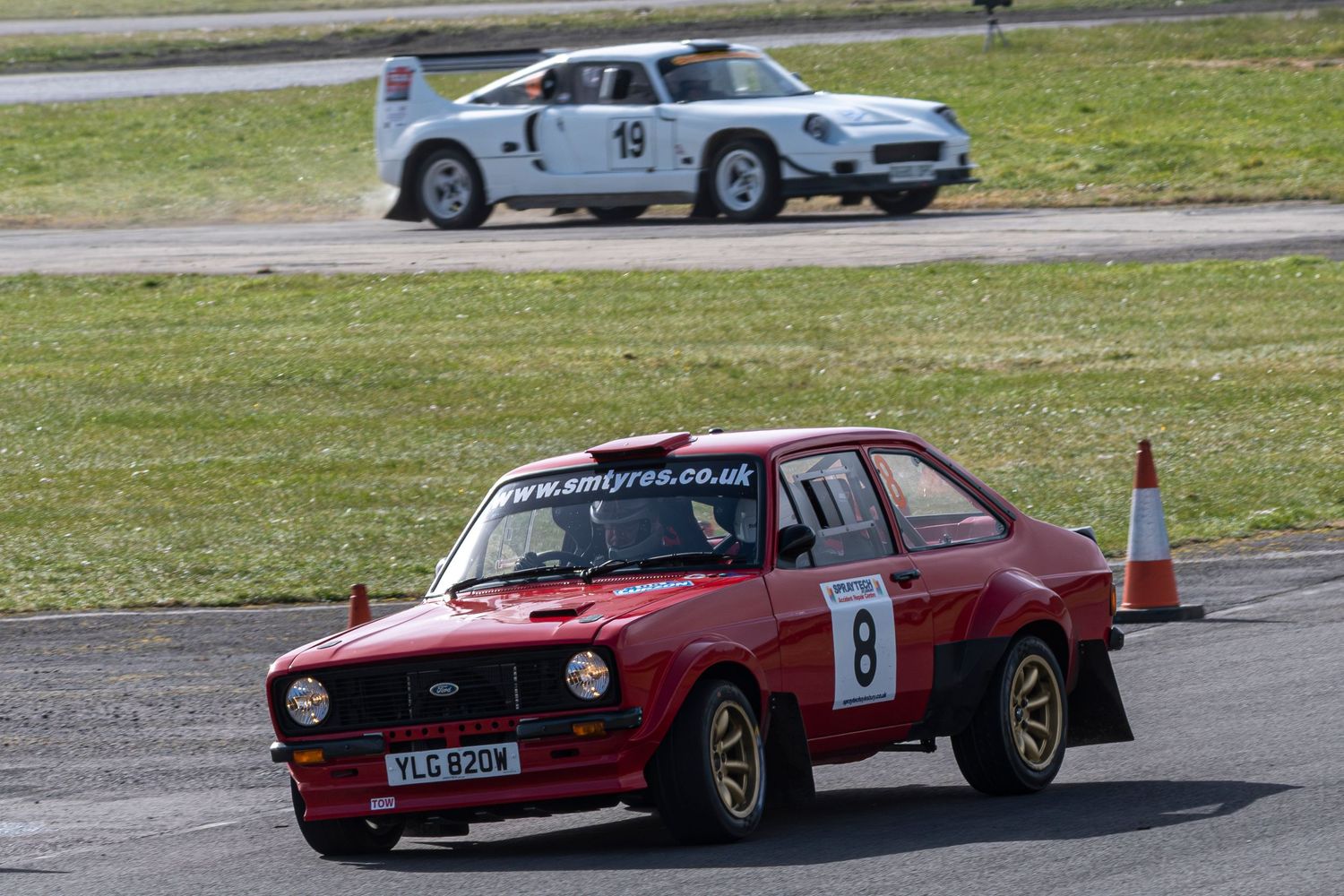 A Ford Escort turns into a corner after another race car shows up in the background.