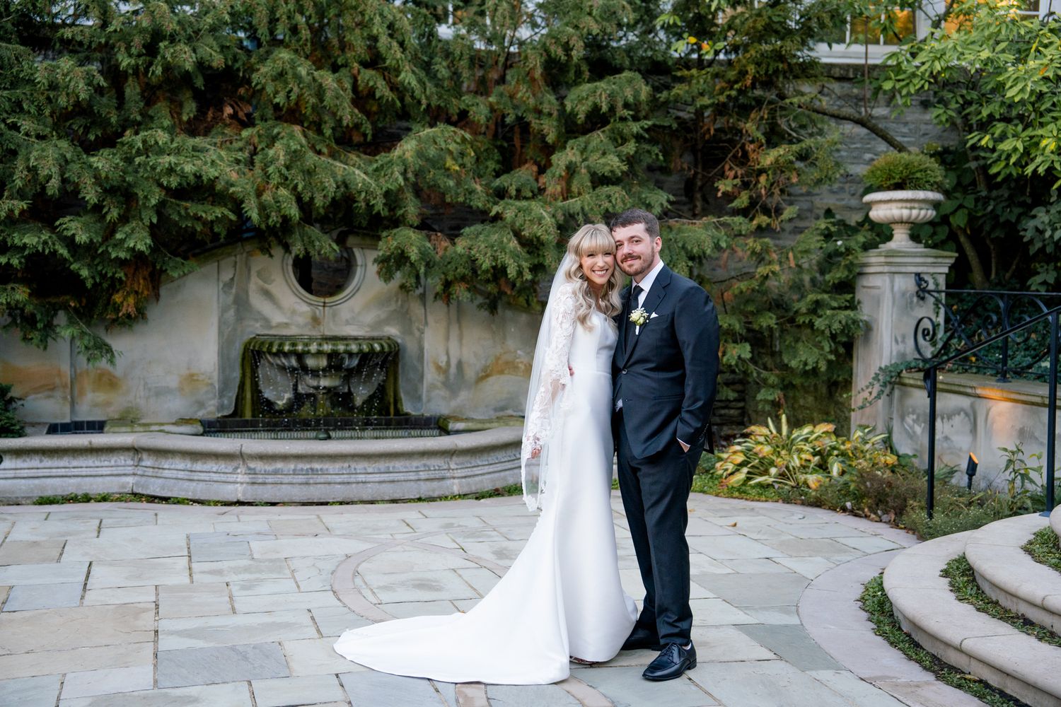 Newly married couple posing together on stone patio with elegant fountain and greenery in background.