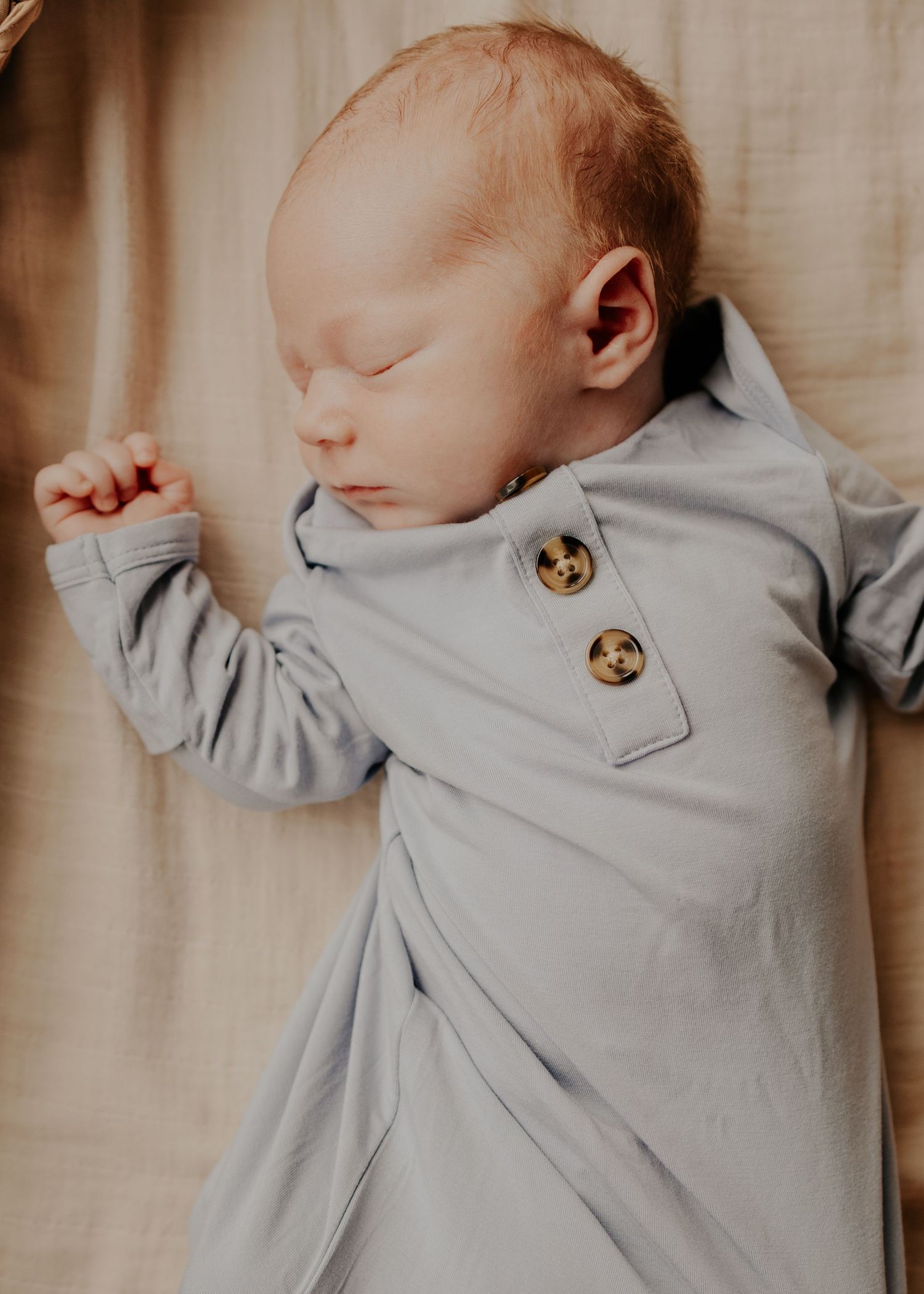 Sleeping newborn baby wearing soft gray button-up outfit while resting on beige fabric background.