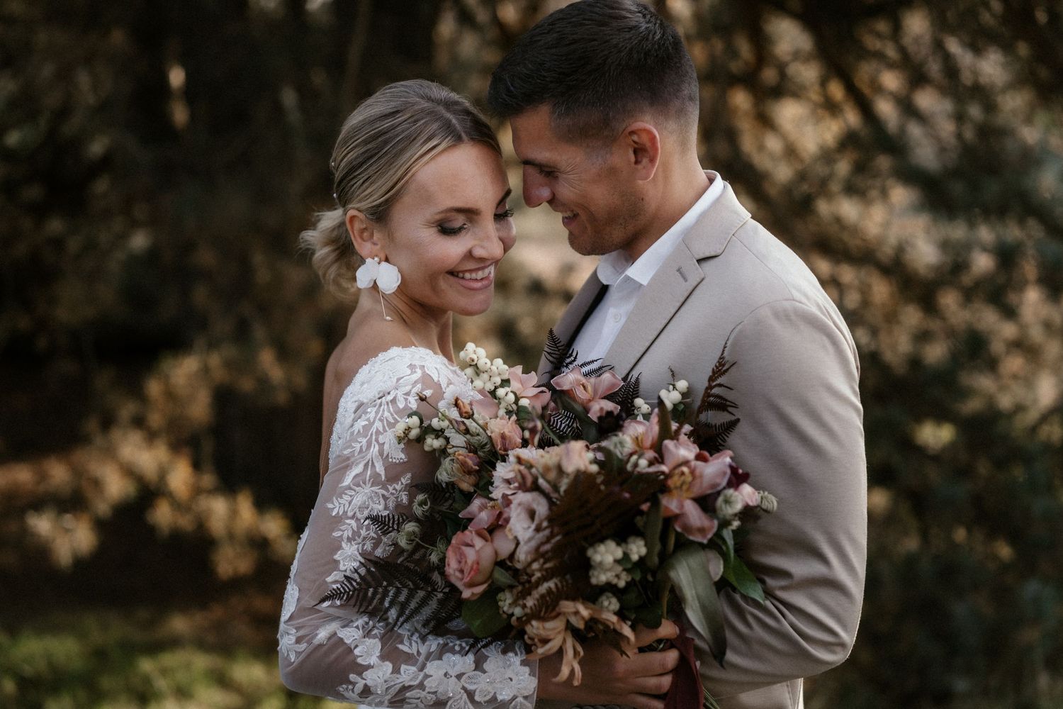 Romantisches Hochzeitspaar umarmt sich und hält dabei einen rosa Blumenstrauß im Freien.