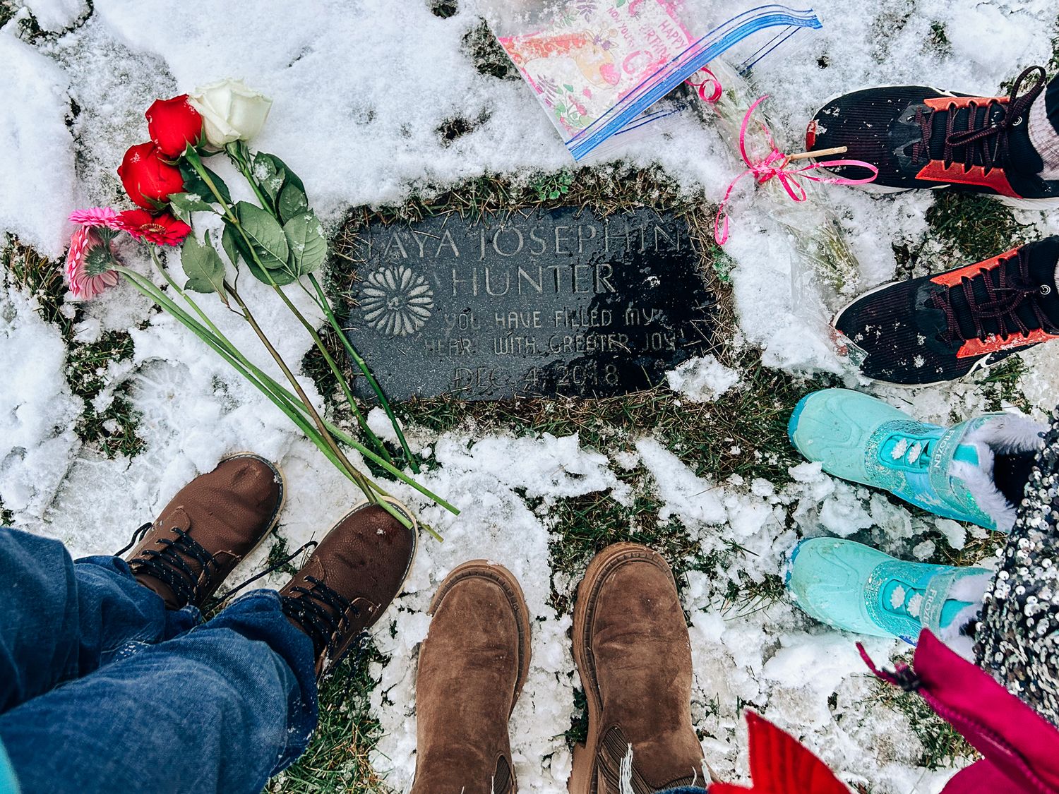 Red roses and winter boots gather around a memorial stone in fresh snow.