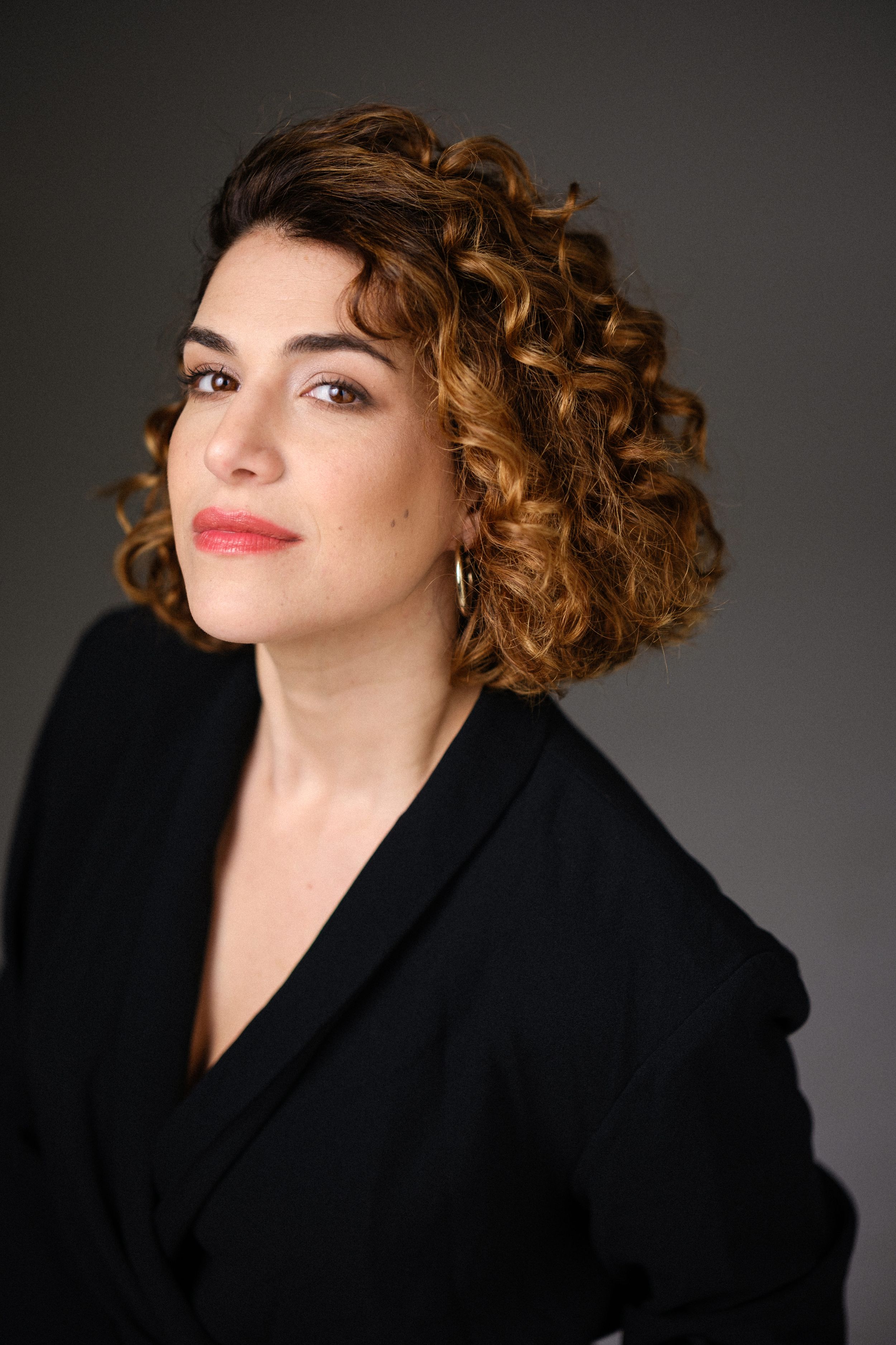 Elegant portrait with styled curls and black blazer against dark background.