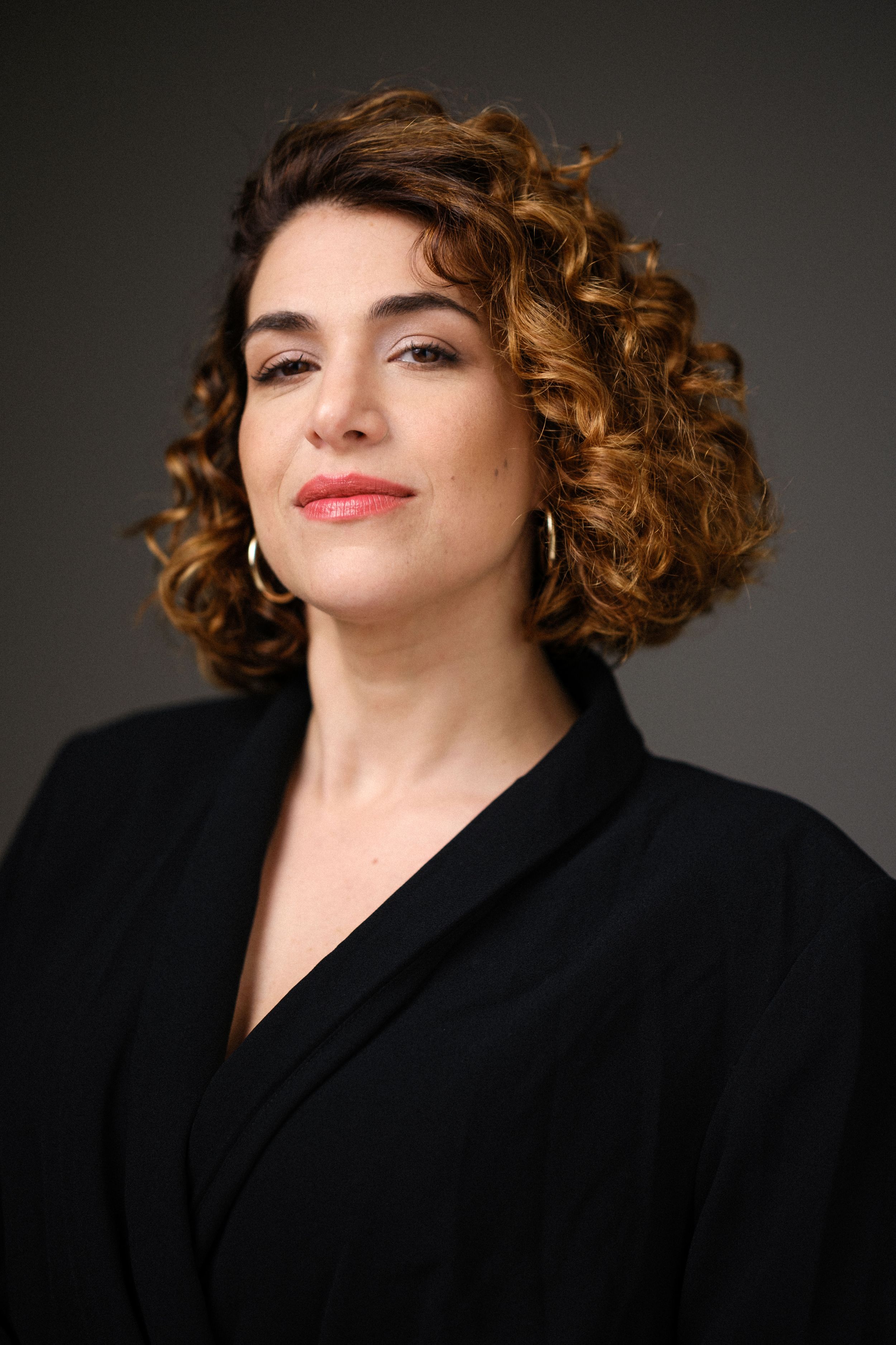 Professional portrait with curly hair and black blazer against dark background showing playful expression.