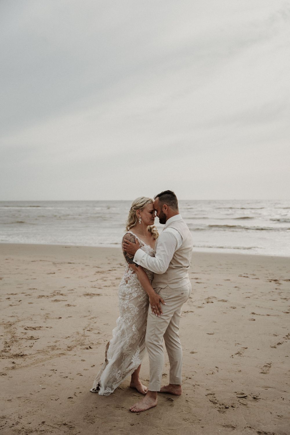 A romantic wedding couple embraces on a misty beach with waves and overcast sky in the background.