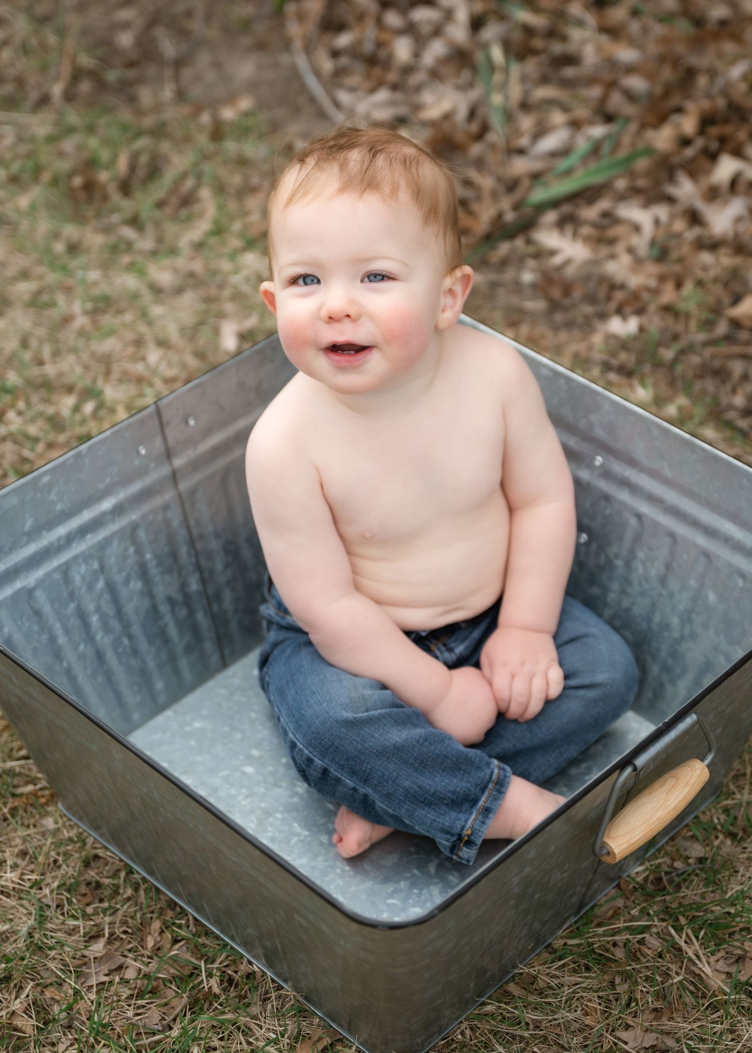 Smiling baby in jeans sitting inside a rustic metal tub outdoors on grass.