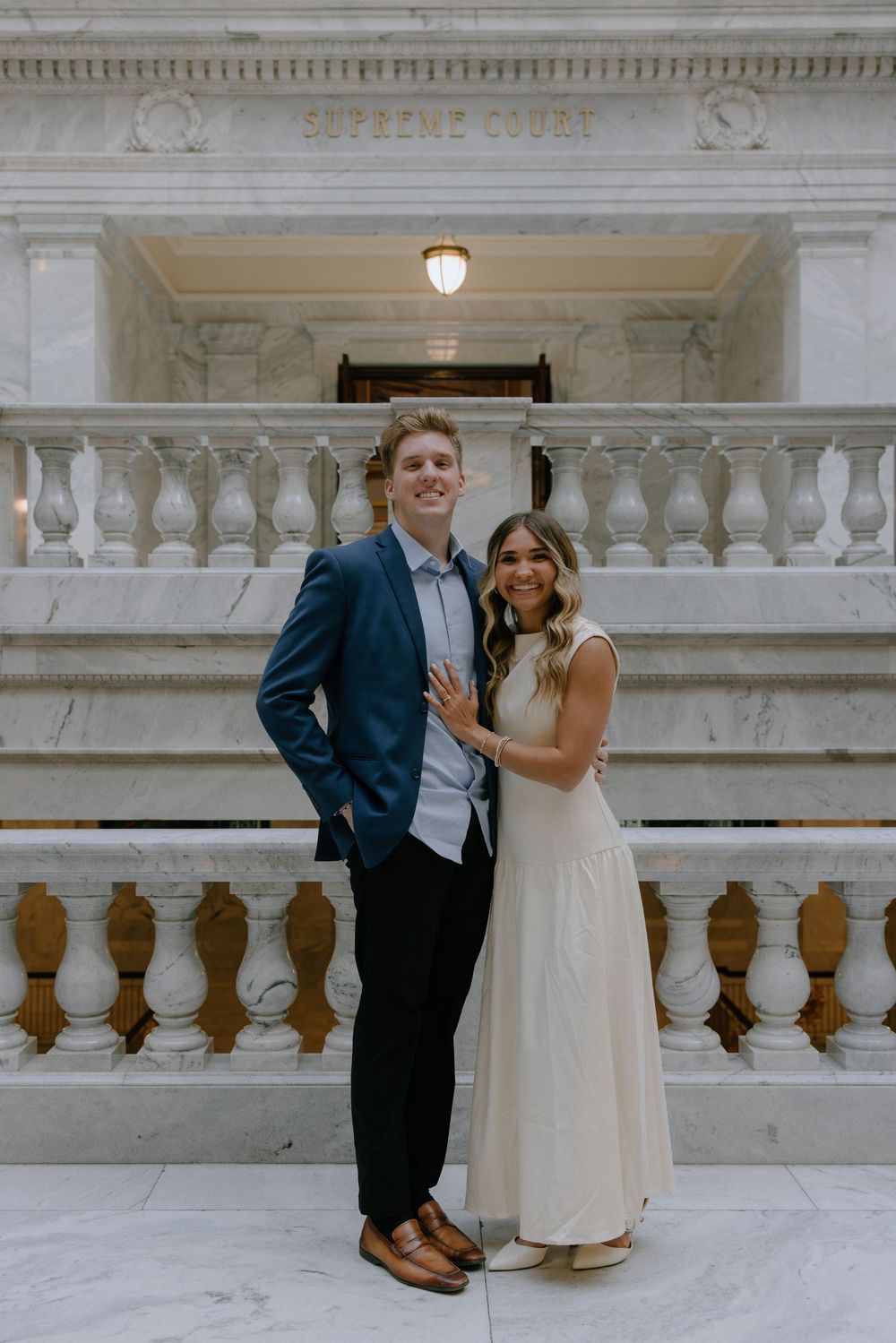 Couple posing on marble steps in front of Supreme Court entrance, man in blue blazer and woman in white dress.