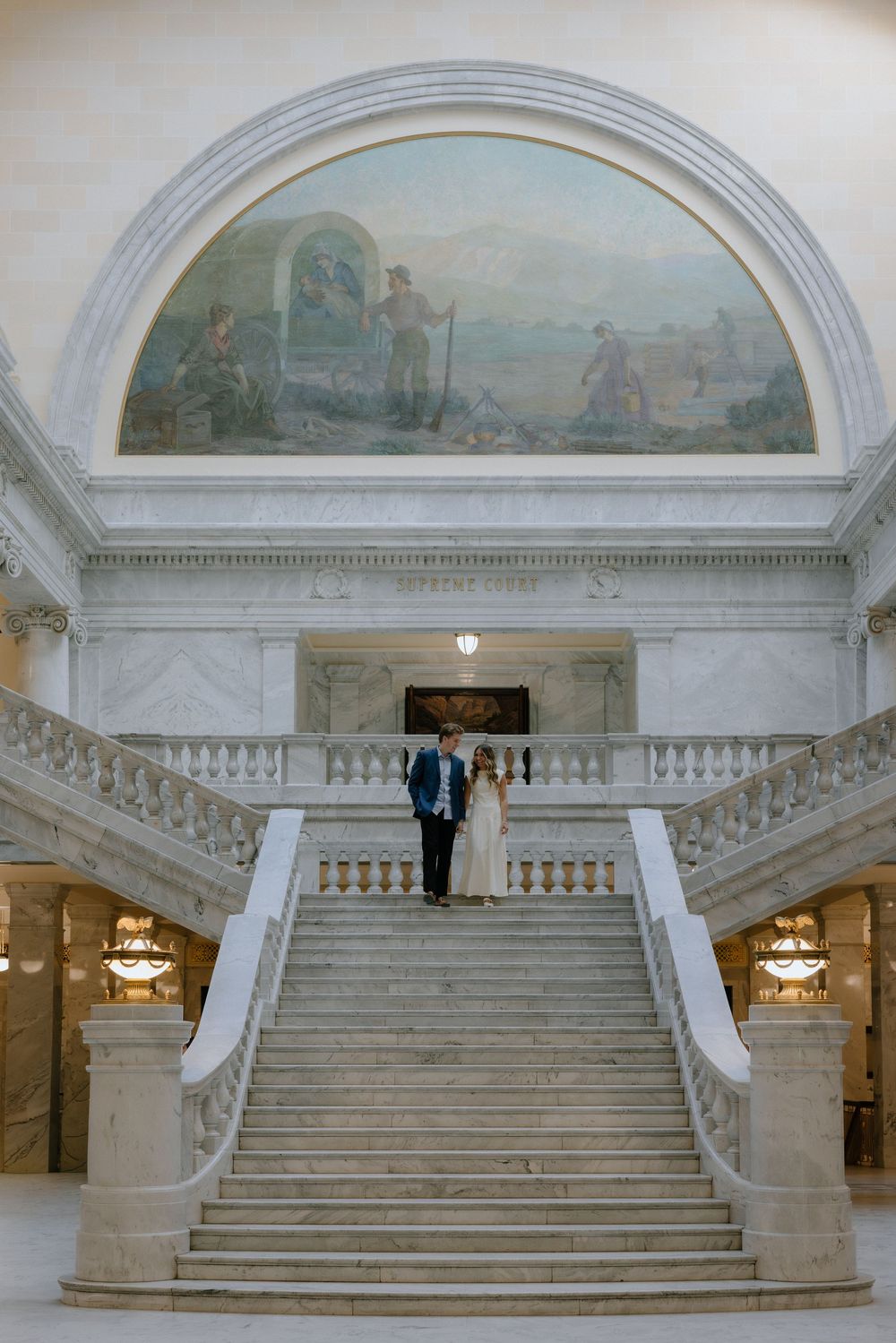 Bride and groom descend grand marble staircase beneath historic mural in elegant capitol building wedding photo.