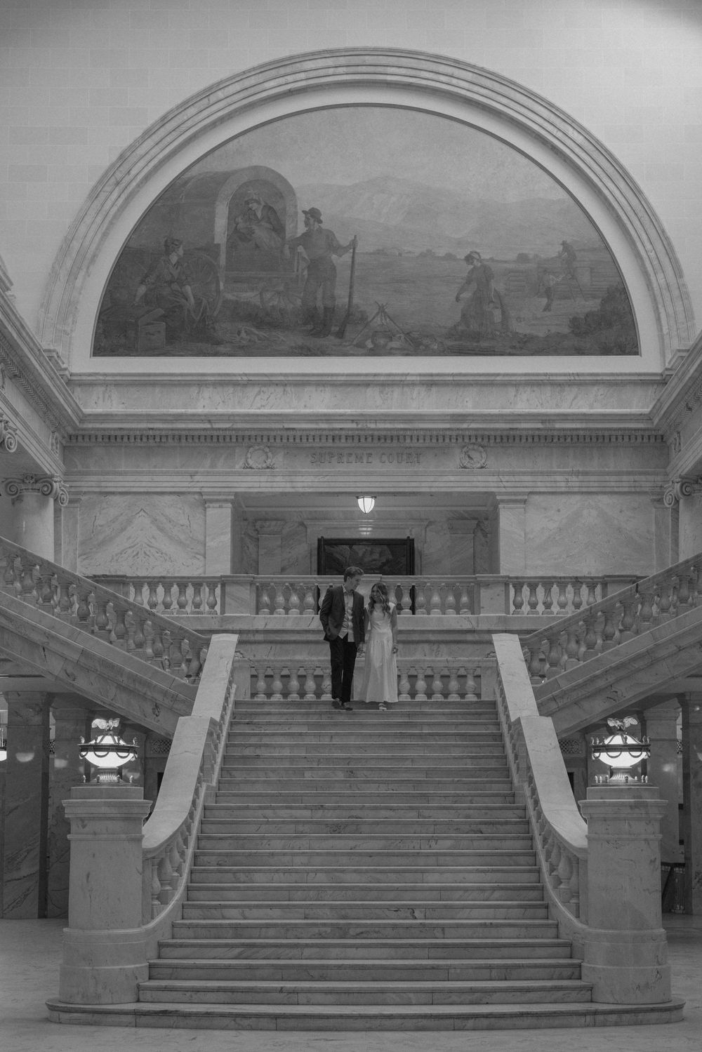Couple poses on grand staircase inside ornate building with mural arch in black and white wedding photo.